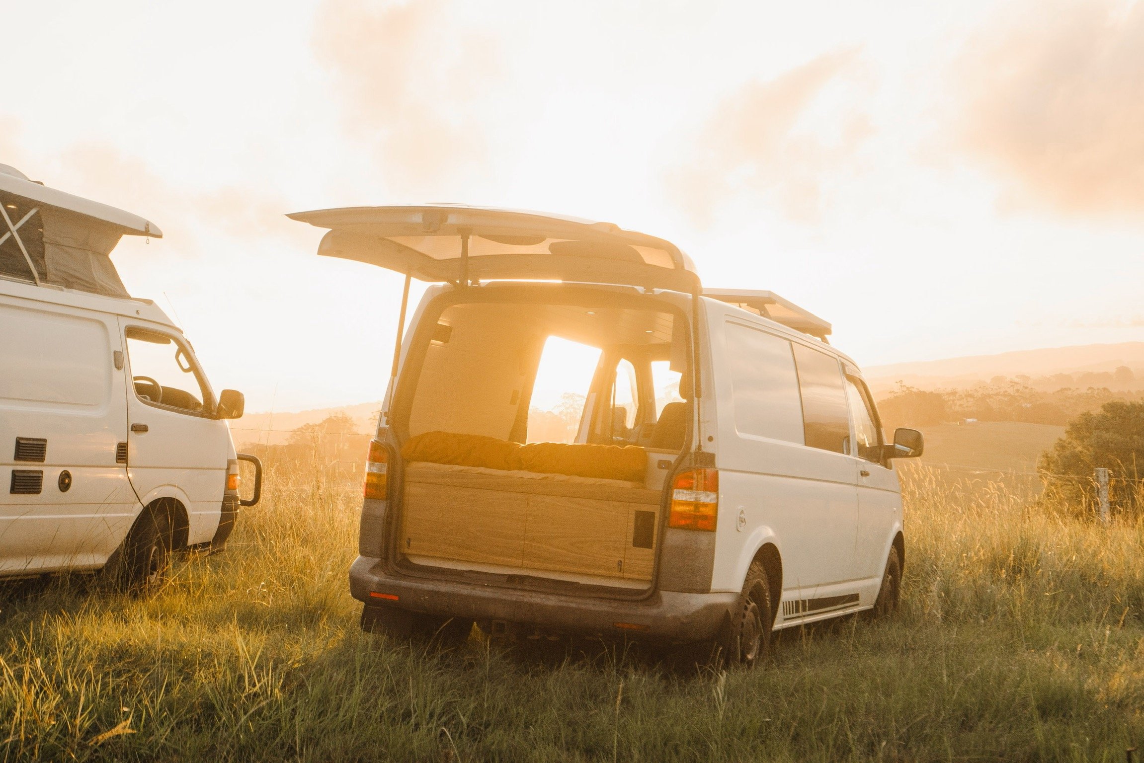 Two white camper vans parked on a grassy field during sunset, with one van's back door open showing an interior bed and the sun shining through the window.