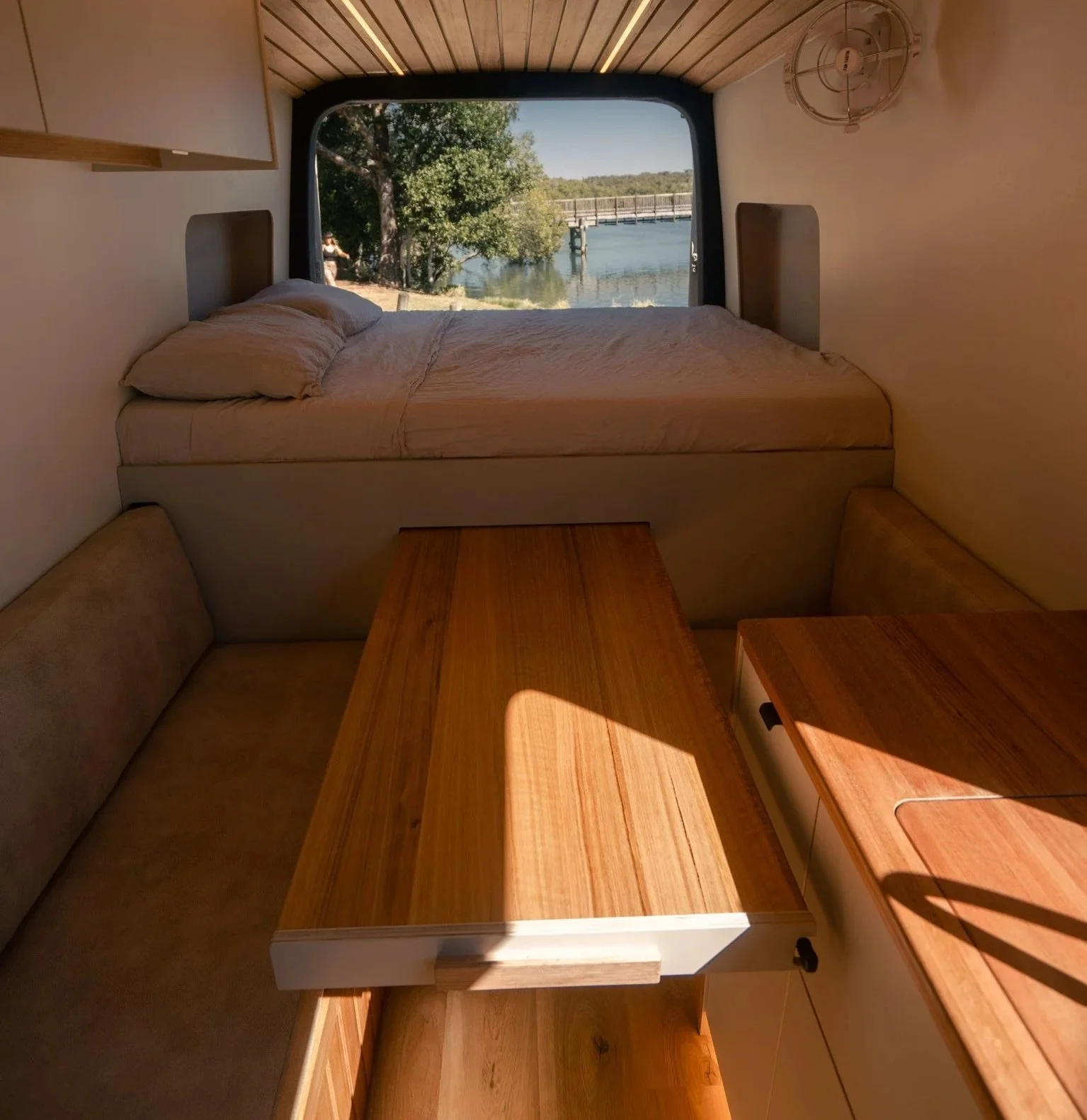 Interior of a cozy camper with a bed near a large window overlooking a river, trees, and a bridge, with wooden furniture and warm lighting.