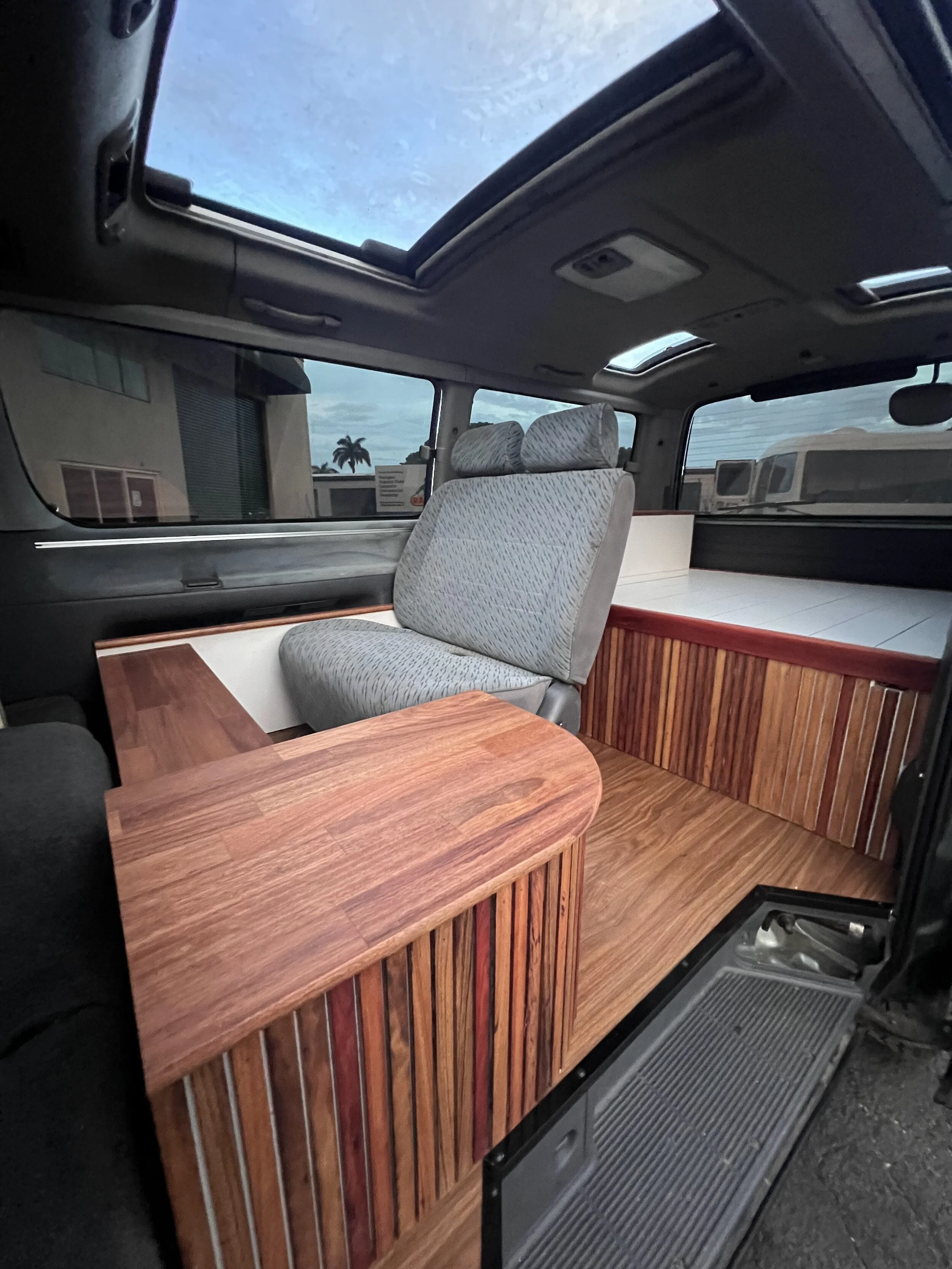 Interior of a van with two gray fabric seats, wooden paneling, a small wooden table, a raised wooden platform, and a large sunroof with a view of the cloudy sky and palm trees outside.