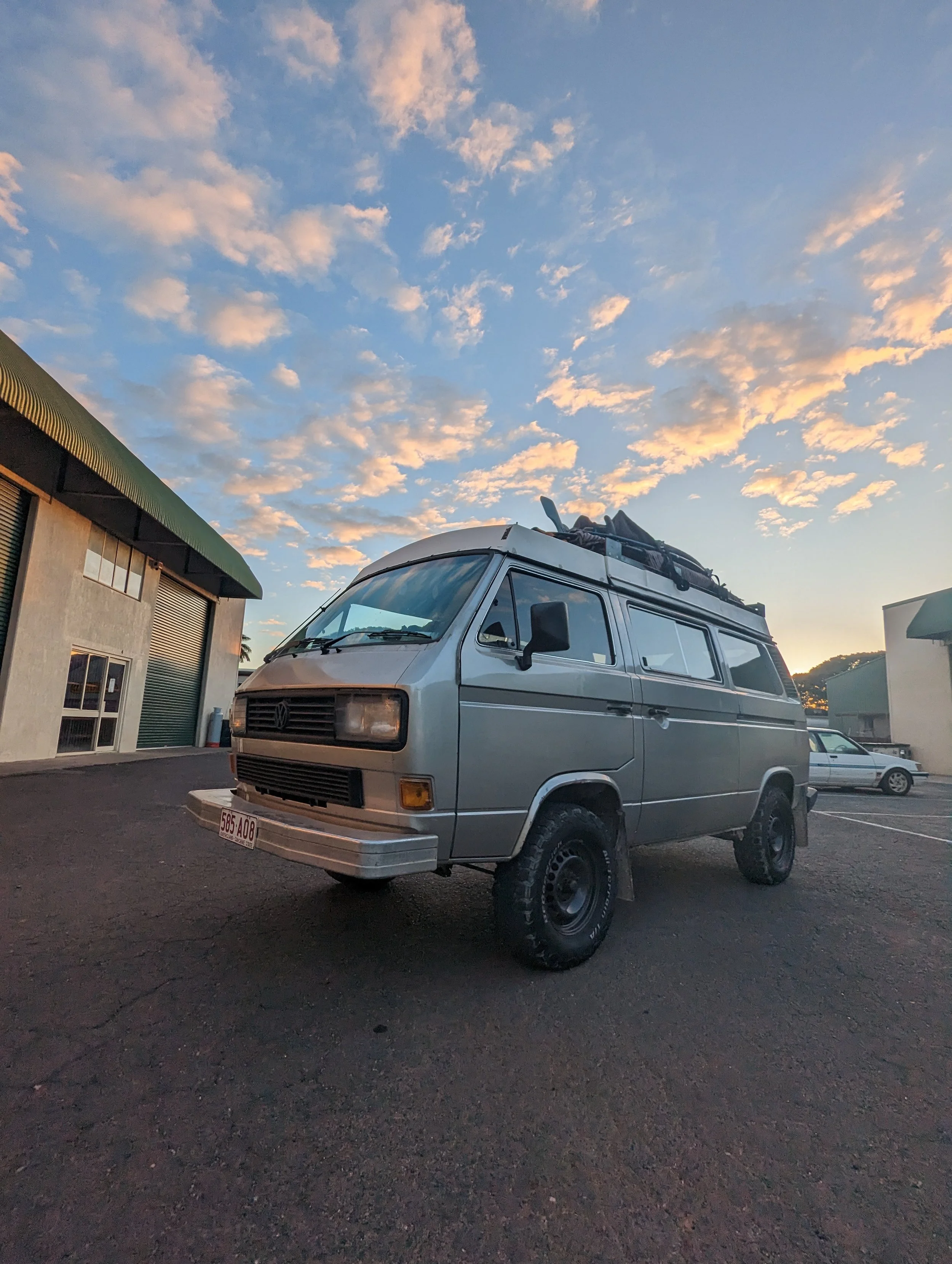 A vintage silver van parked on a paved lot at sunset with a dirty sky filled with scattered clouds, surrounded by industrial buildings.