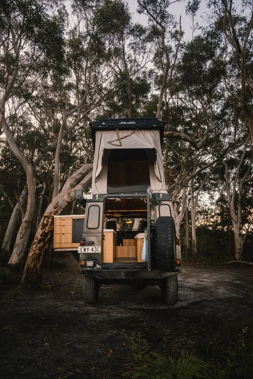 A camper vehicle parked in a wooded area with large trees around it, showing an open rear door revealing a small interior with wooden furniture.