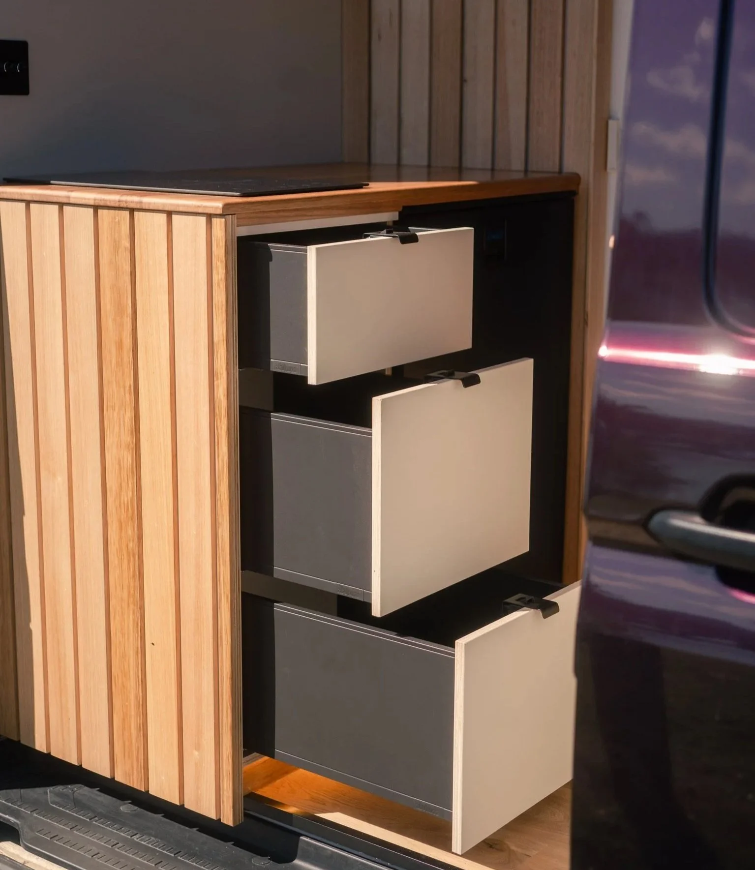 Open drawer cabinet with three pull-out drawers in a wooden cabinet, set outdoors near a parked vehicle.