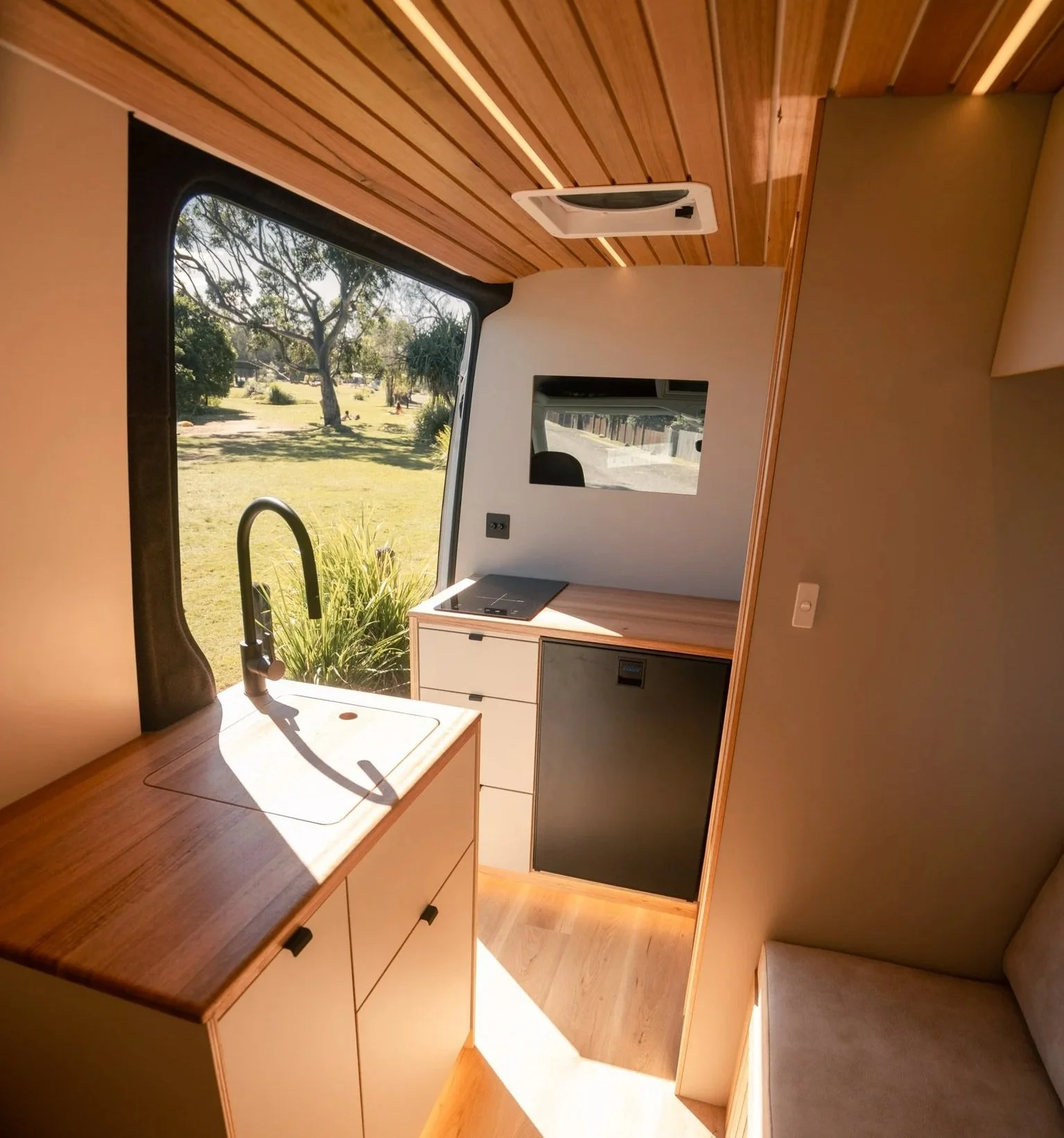 Small kitchen area inside a tiny home with a window view of trees and grass, a black sink with a black faucet, a wooden countertop, a small black fridge, and a stove. There's a mirror and window in the background and wood-paneled ceiling.