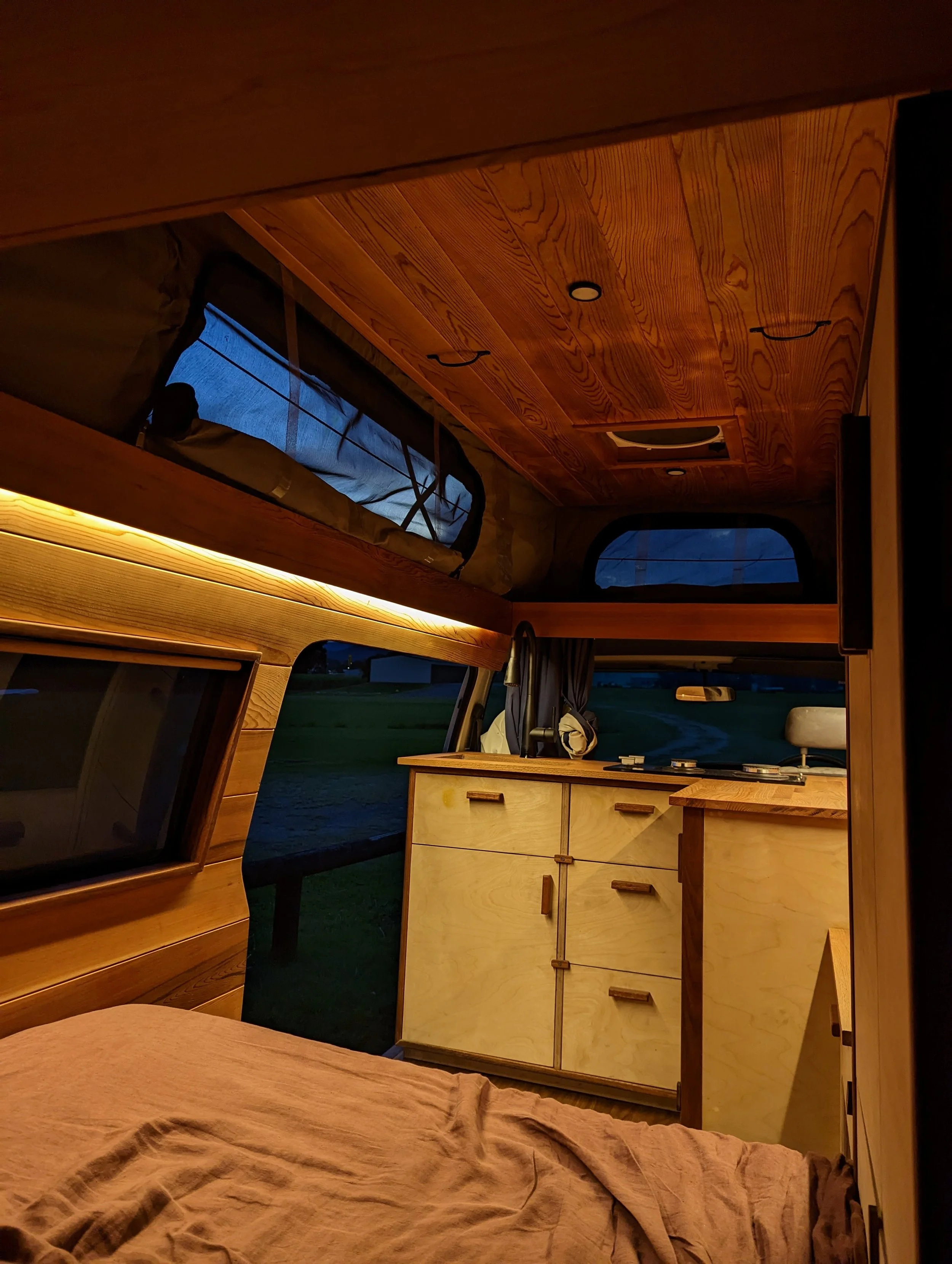 Interior of a camper van with wooden paneling, bed, small cabinet, windows, and subtle lighting at dusk.