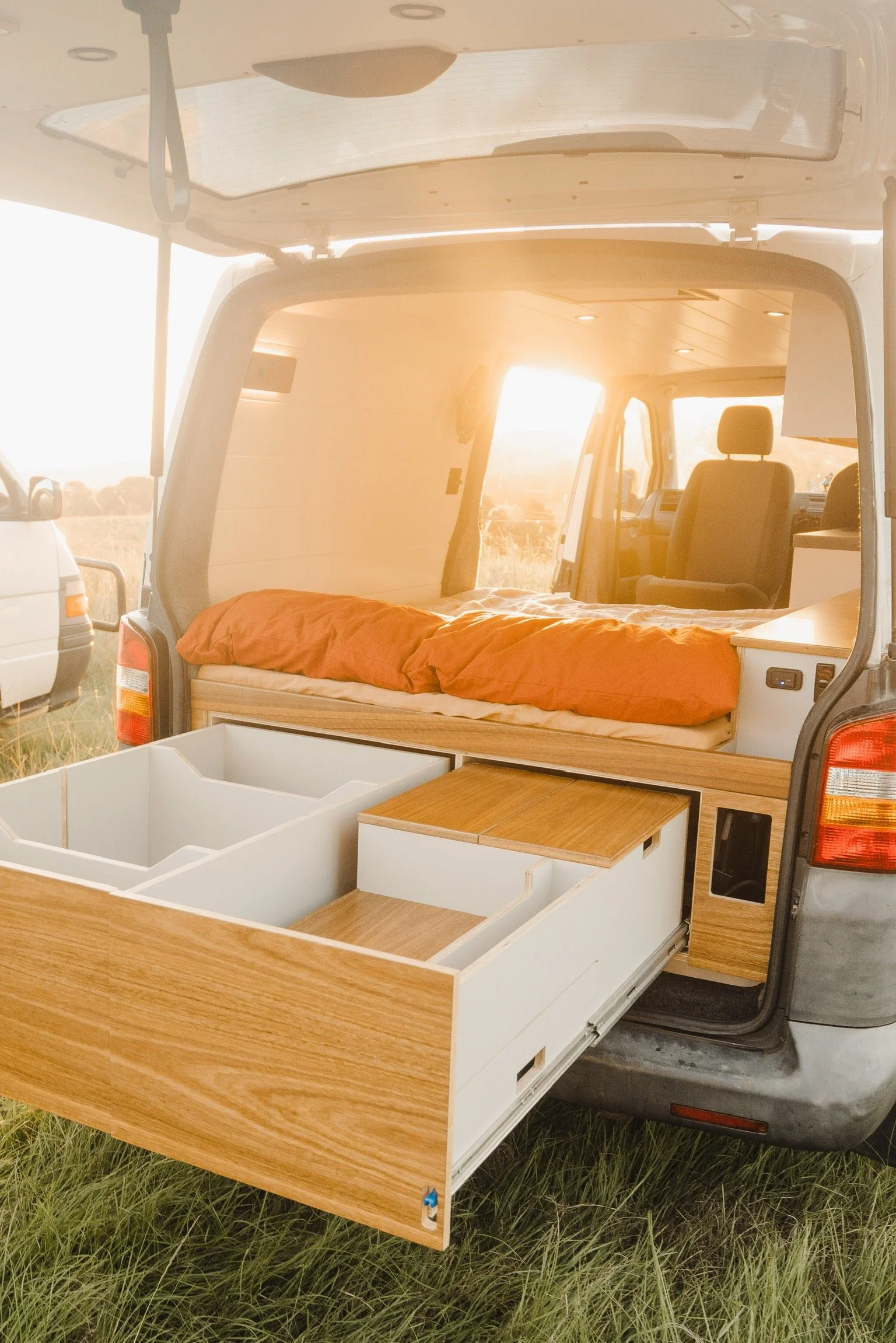 The interior of a camper van with an open rear hatch, showing a bed with orange bedding, wood and white storage compartments, and a view of the dashboard and driver seat through the front window at sunset.