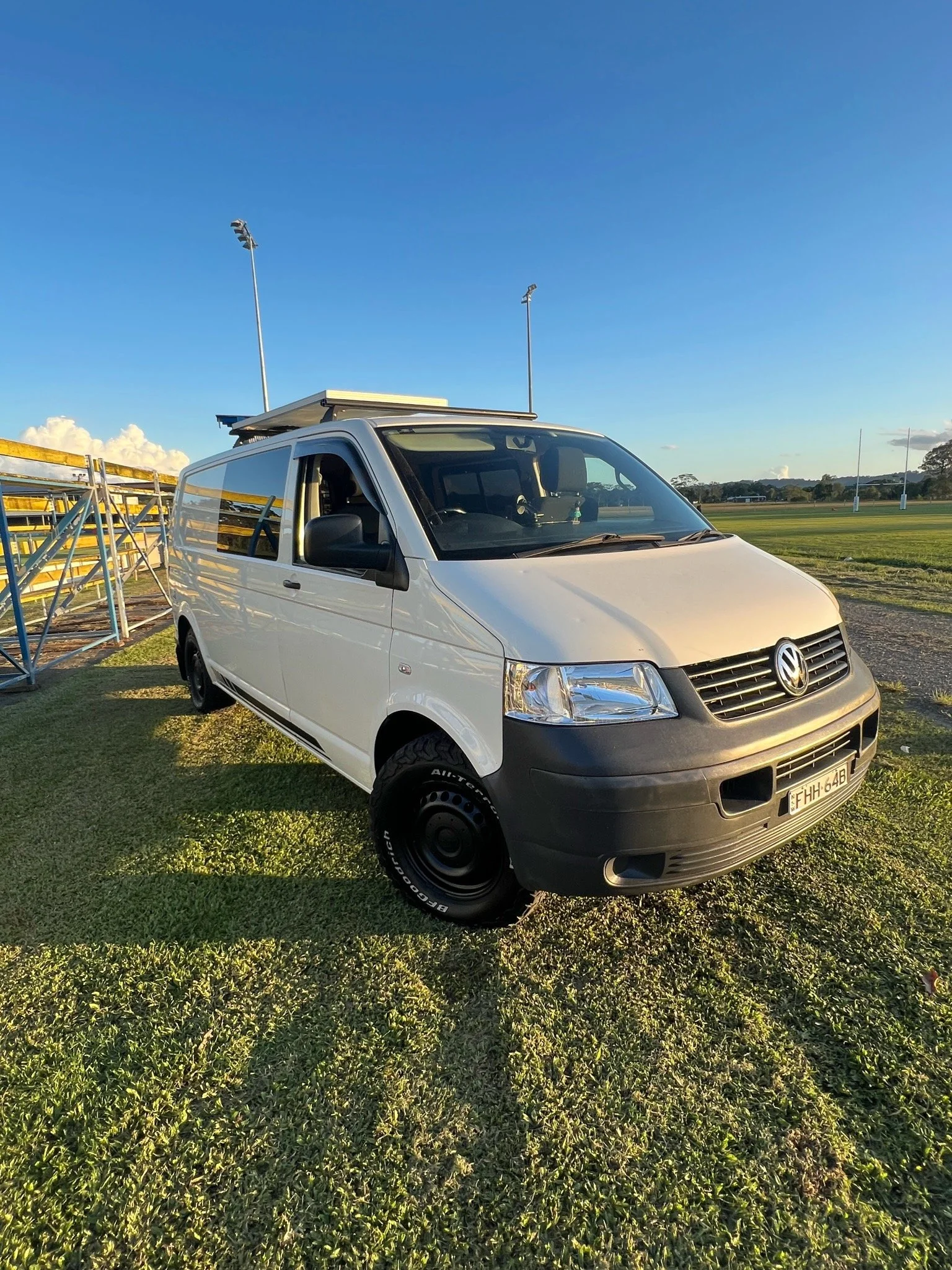 A white Volkswagen van parked on a grassy field with stadium lights and a sports field in the background under a clear blue sky.