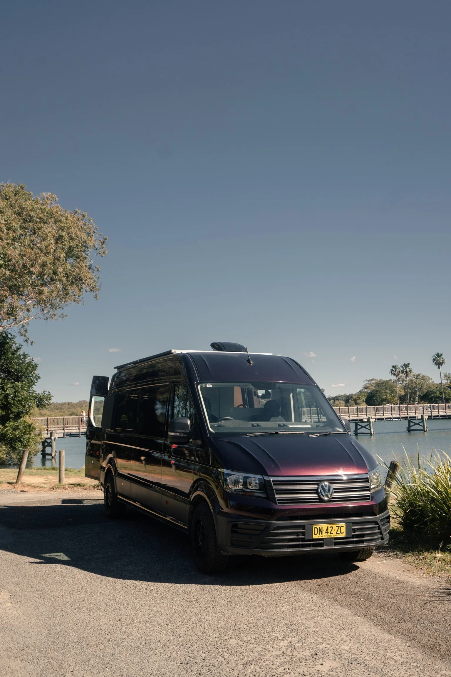A black van parked on a road near a body of water, with a bridge and trees in the background on a clear day.