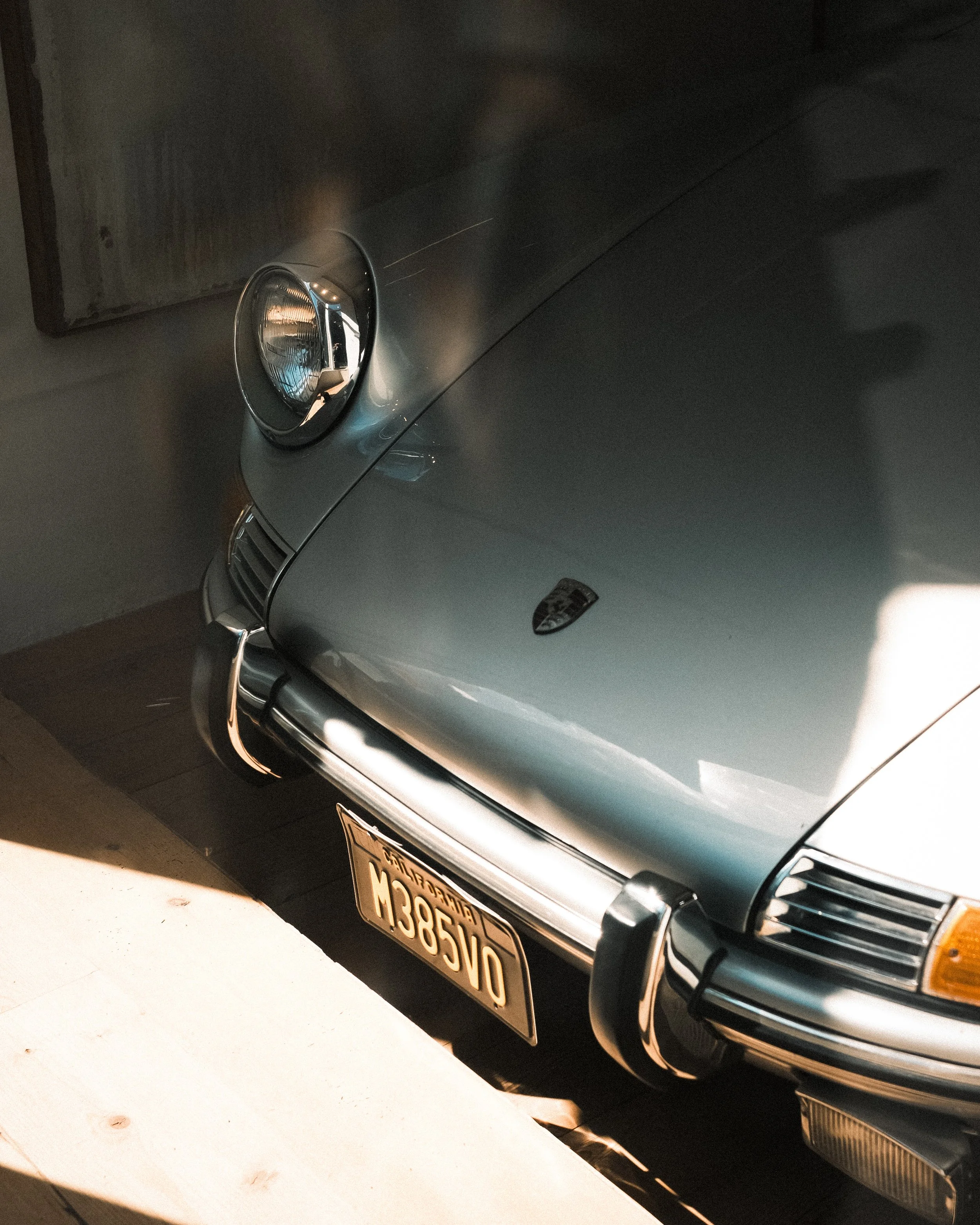 Close-up of the front part of a gray Porsche sports car, showing its headlight, grill, bumper, and a California license plate with the characters H385VB.