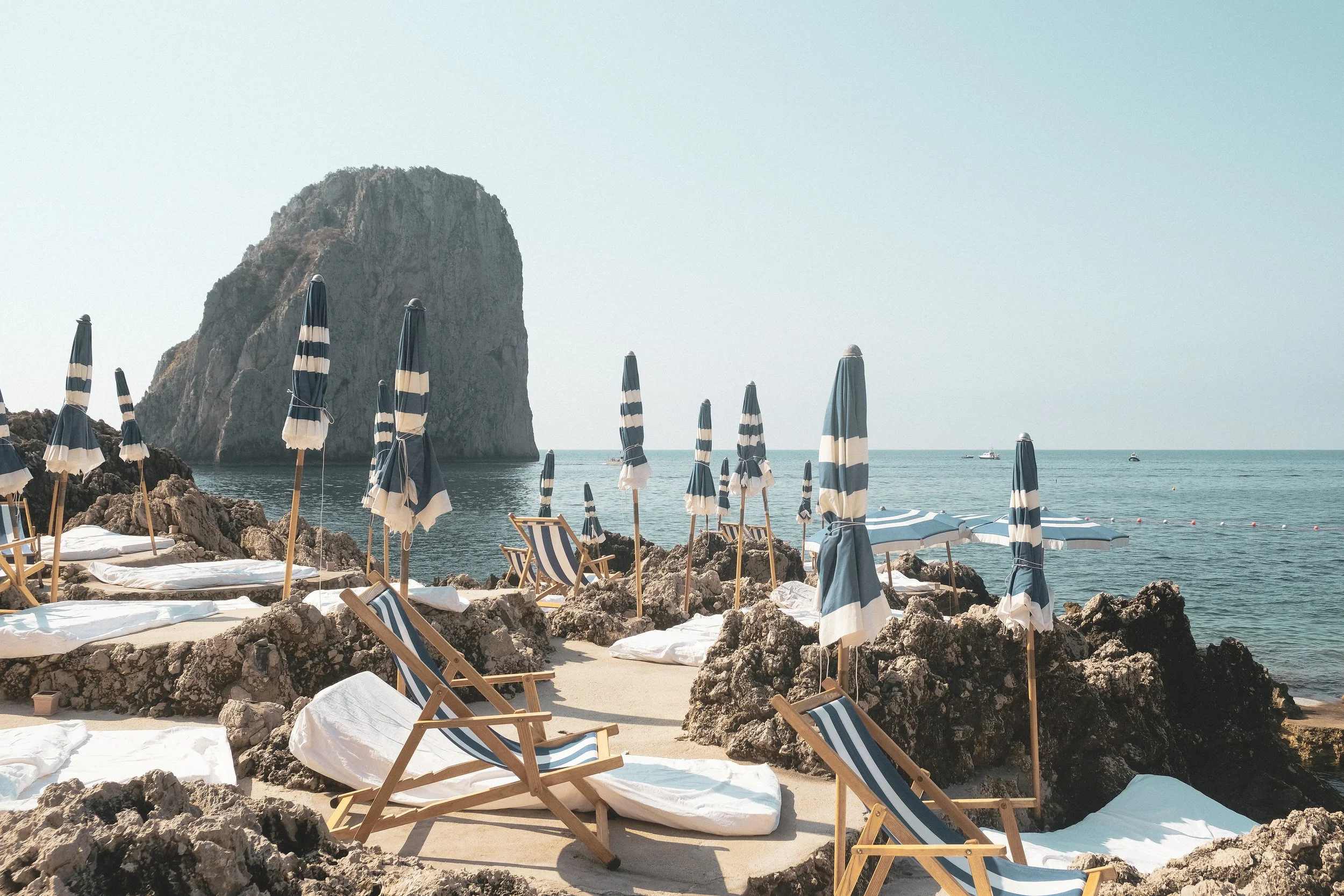 Beach with striped umbrellas and lounge chairs on rocky shore with large sea stack in background and boats on the ocean