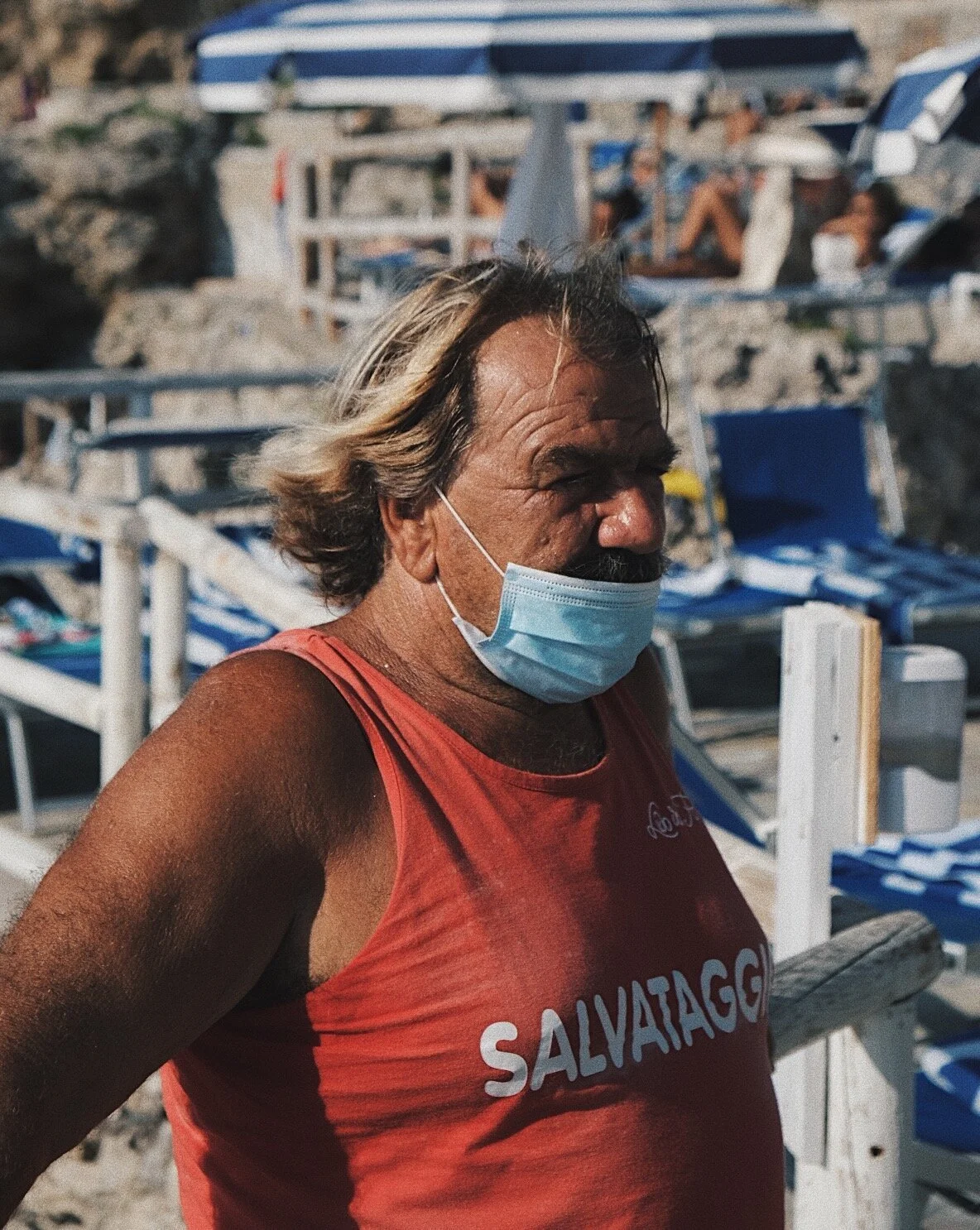 A man with long gray hair wearing a red sleeveless shirt with the word 'SALVATAGG' on it, and a face mask, standing on a beach with lounge chairs and umbrellas in the background.
