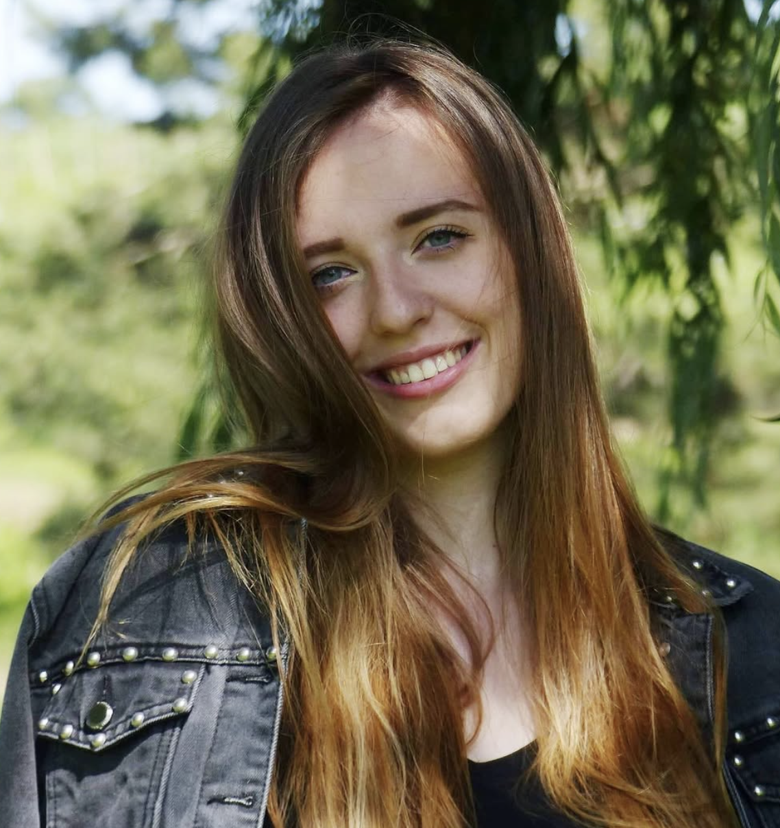 A young woman with long red hair and blue eyes smiling outdoors, wearing a black top and a dark jacket with studs, surrounded by green foliage.