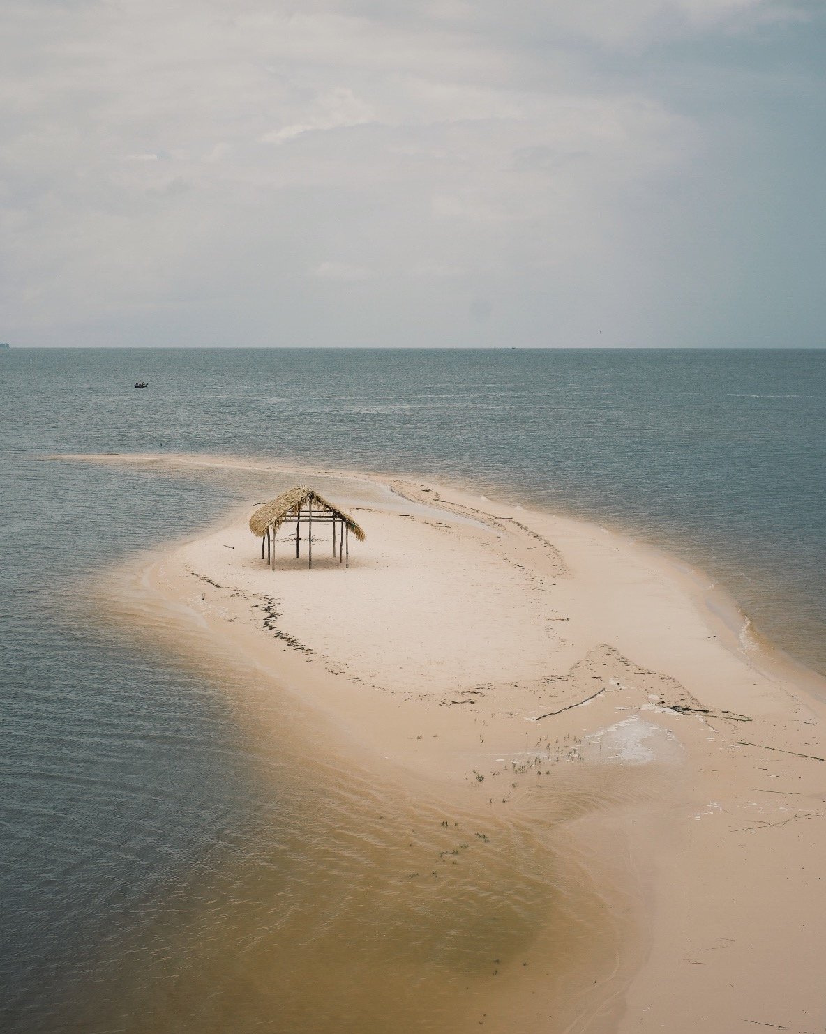 A sandy beach with a small thatched-roof shelter on a sandbar in the ocean, with boat in the distance under cloudy sky.