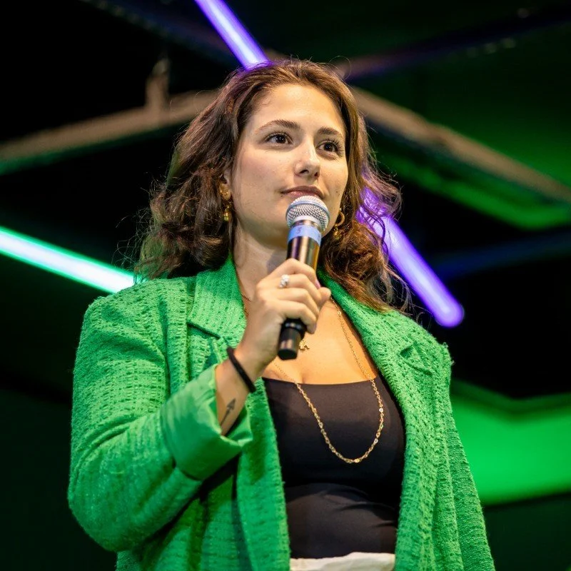 Woman with wavy brown hair wearing a green textured blazer and black top, holding a microphone and speaking on stage with colorful lights in the background.