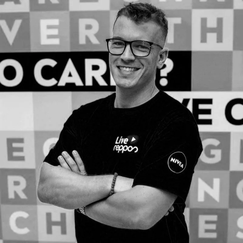 Young man smiling, wearing glasses and a black t-shirt with 'Live reppo' and 'NIVEA' logos, standing with arms crossed in front of a patterned wall with text.
