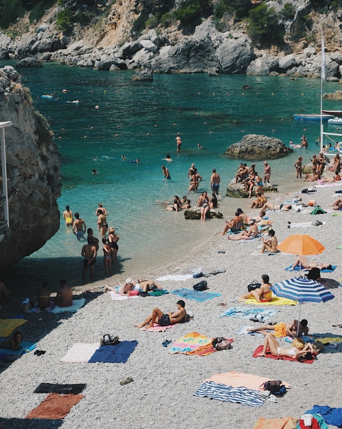A crowded beach with people sunbathing, swimming, and socializing near the rocks and turquoise water, with umbrellas and beach towels scattered on the pebbled shore.
