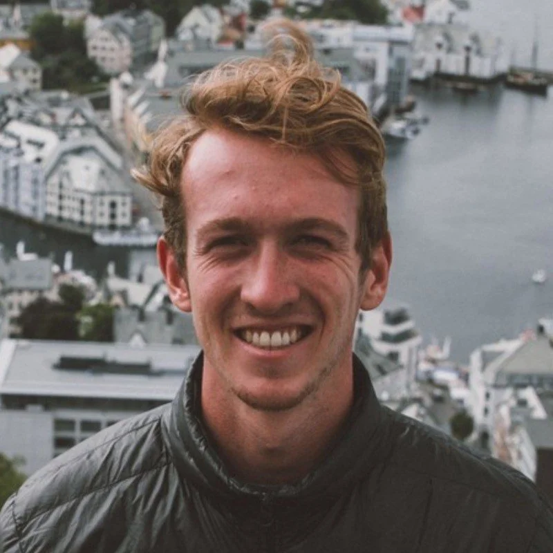 Young man with wavy brown hair smiling outdoors with a city and water in the background.