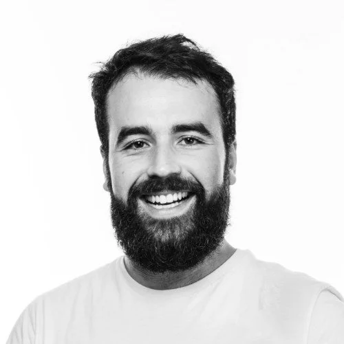 Portrait of a smiling man with a beard and short hair, wearing a light-colored T-shirt, against a plain white background.