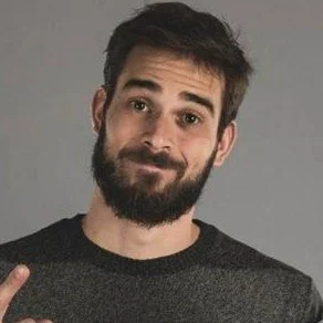 A young man with dark hair and a beard wearing a dark gray shirt, smiling subtly against a gray background.