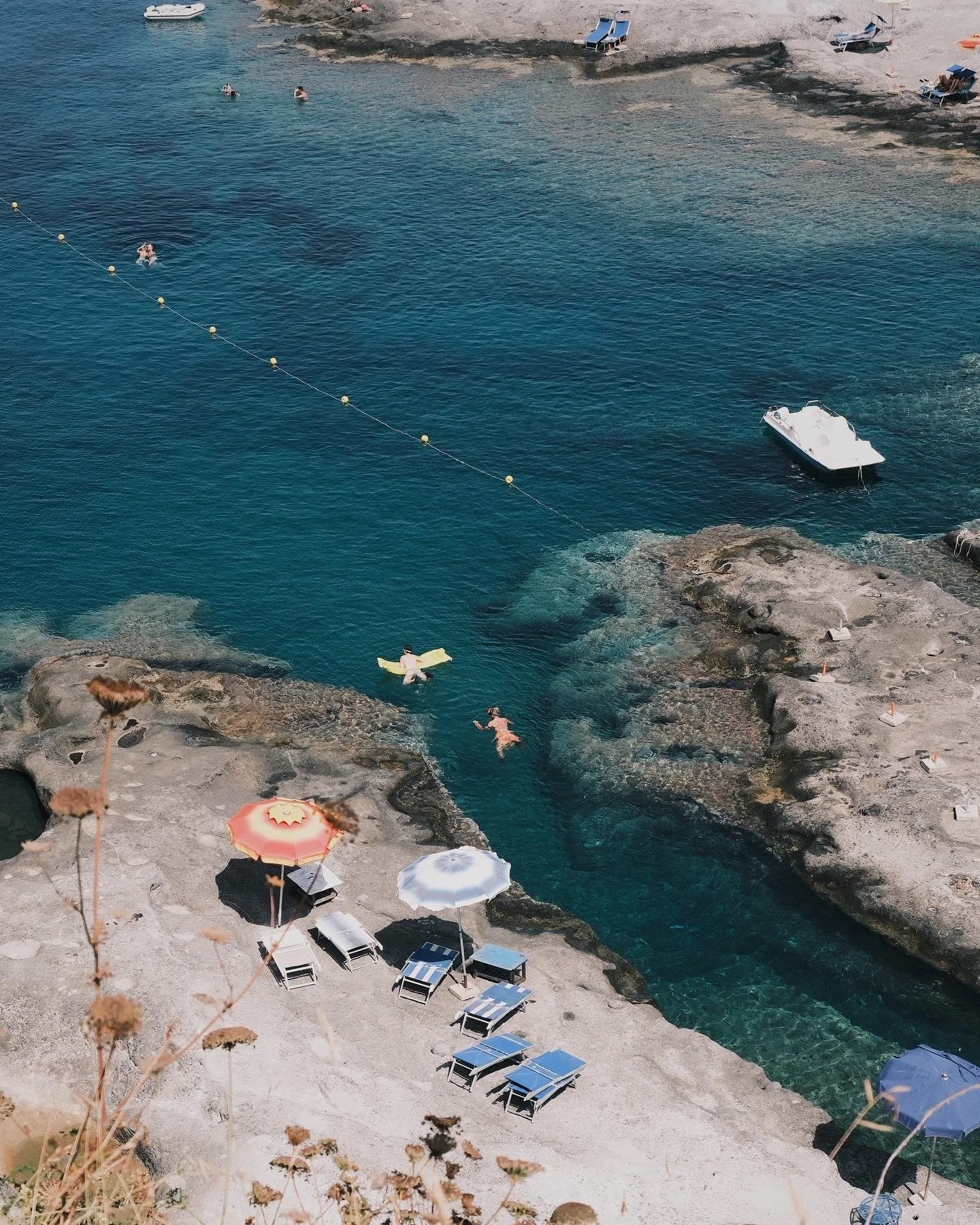 People swimming and relaxing on a rocky beach with umbrellas and lounge chairs, clear blue water, and boats nearby.