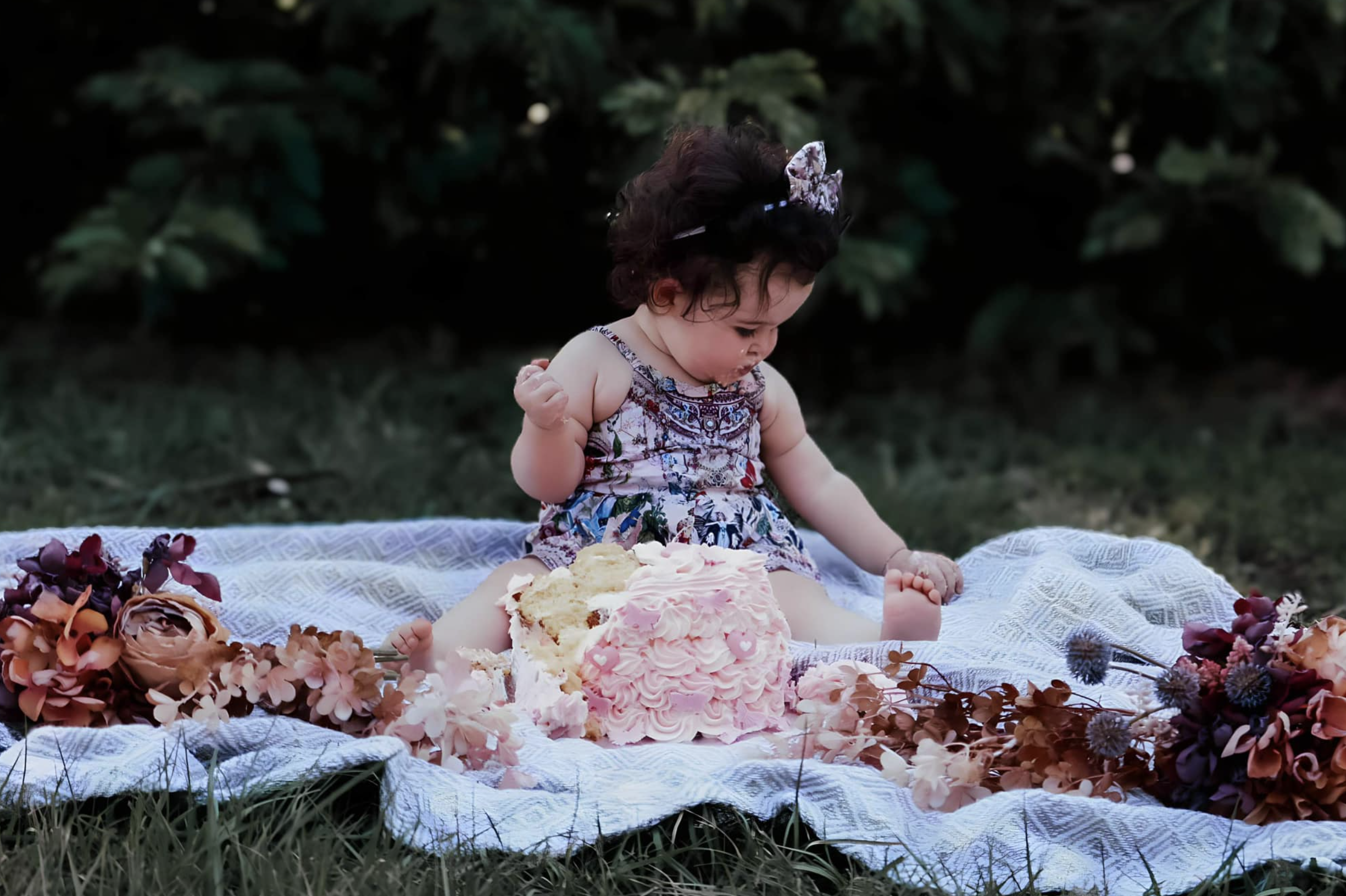 A young girl sitting on a blanket outdoors with a decorated cake and flowers.