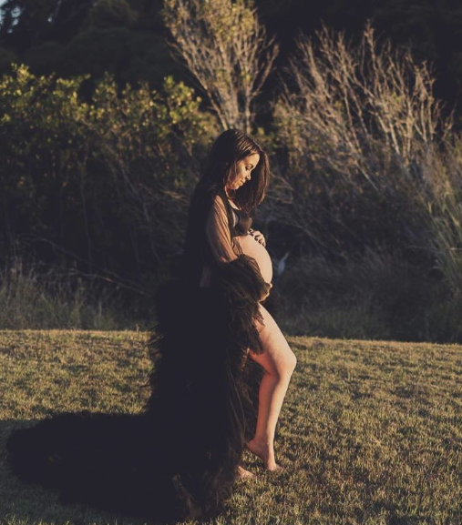 A pregnant woman is outside on grass, with a woman kneeling in front of her, seemingly embracing her belly. There are trees in the background, and the scene appears to be during sunset or sunrise.