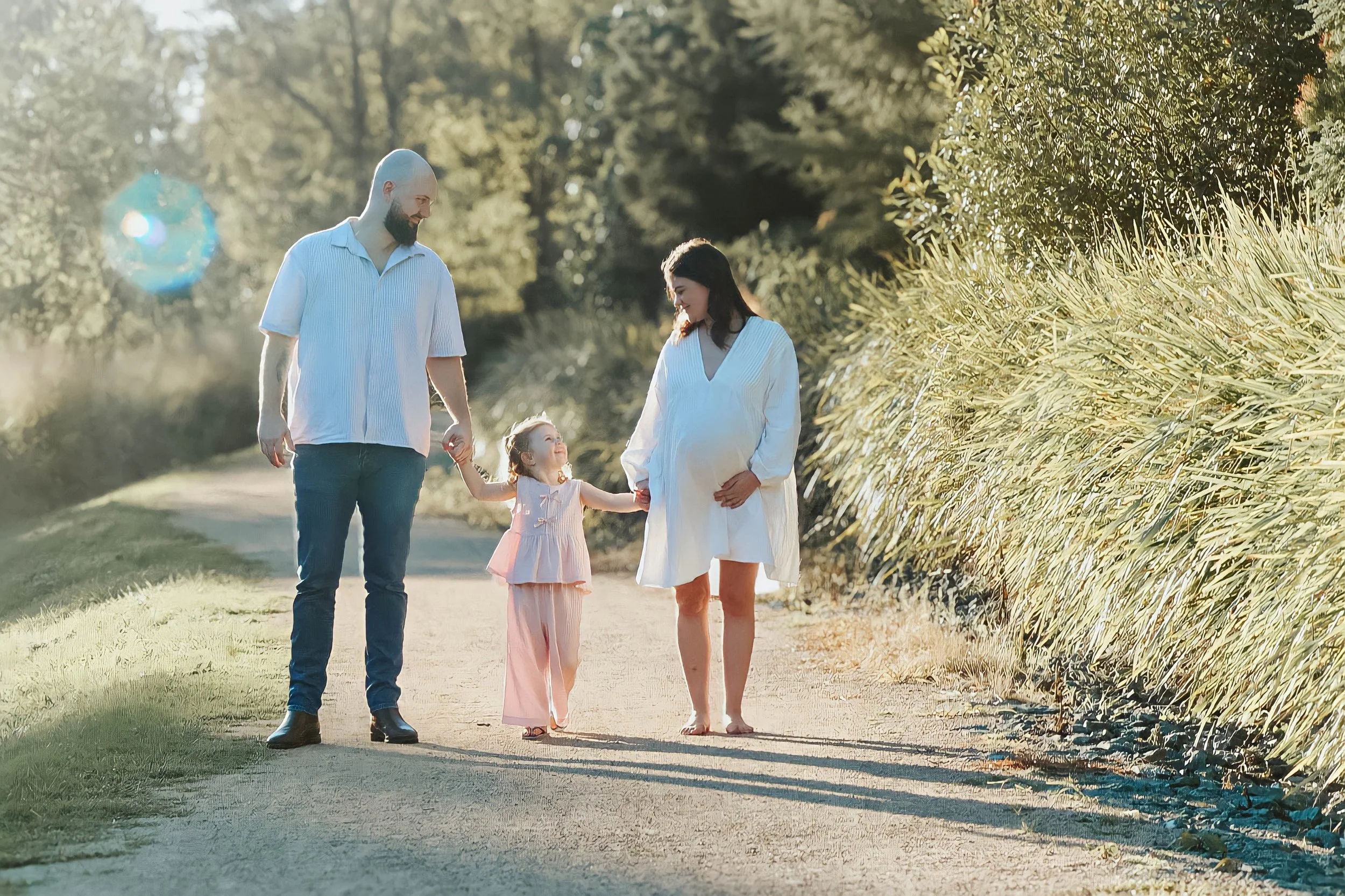 A family of three walking outdoors during the daytime. The man is holding the child's hand, and the woman is pregnant, holding her belly and walking barefoot. The setting appears to be a rural or park area with trees and tall grass.