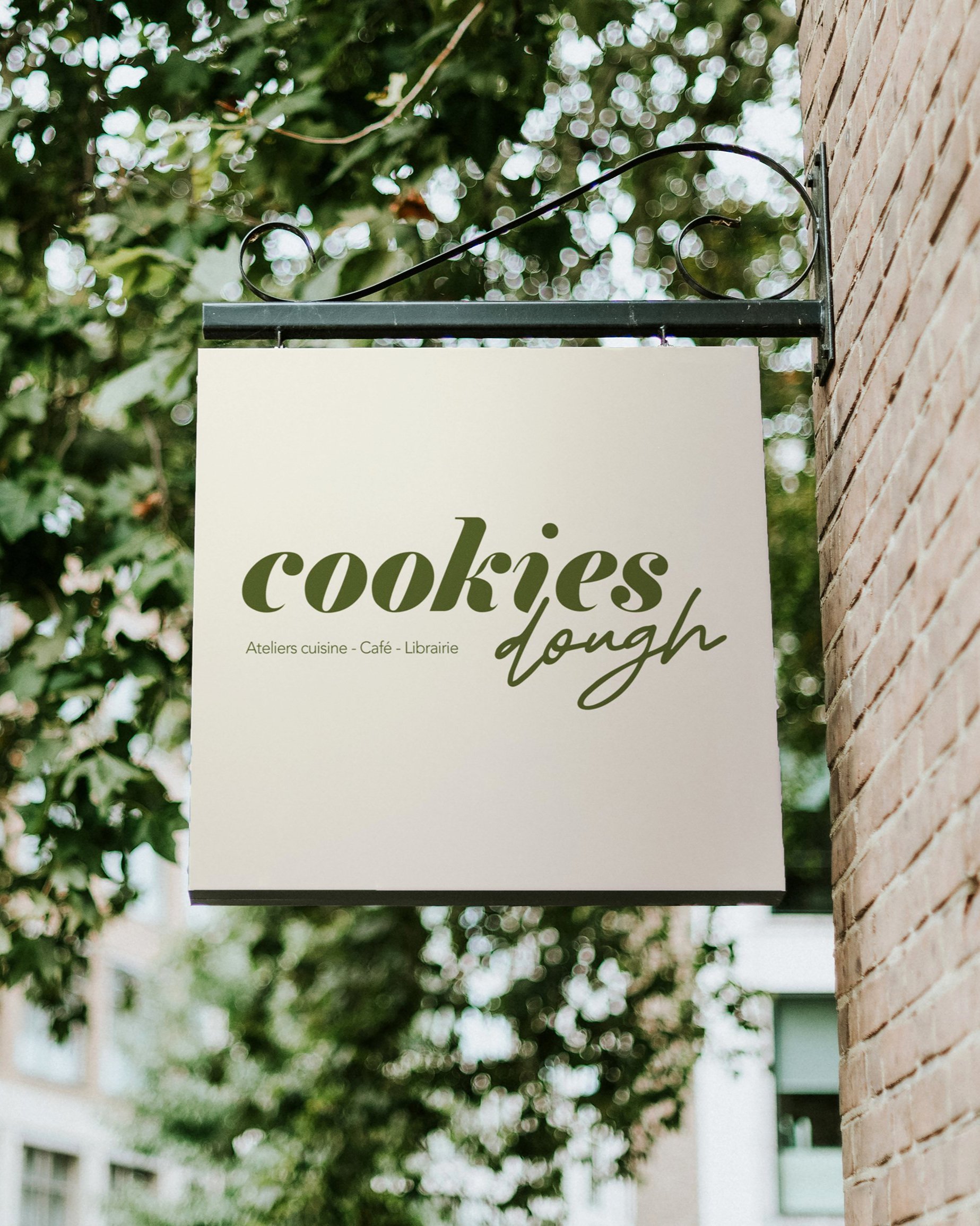 Panneau blanc avec écrit en vert 'cookies dough' et en petits caractères 'Ateliers cuisine - Café - Librairie', fixé à un mur en brique.