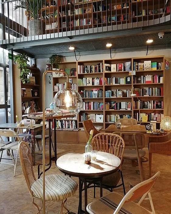 Une bibliothèque ou librairie avec des étagères remplies de livres, un mobilier en bois, et des lampes, dans une ambiance chaleureuse et cosy.
