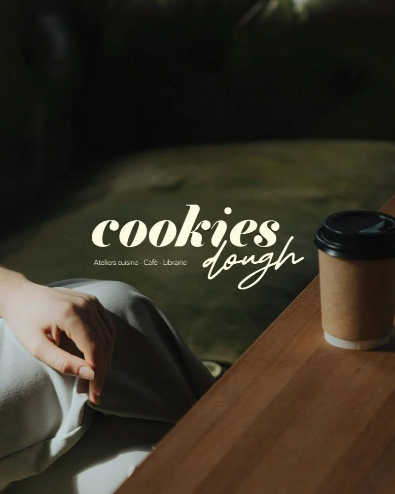 Une tasse à café en carton sur une table en bois à côté d'une main posée sur une jambe, fond sombre, texte en superposition : "cookies dough" avec des sous-titres : ateliers cuisine, café, librairie.