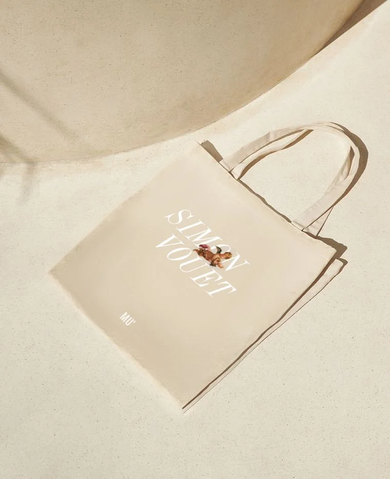 Un sac en toile beige avec le logo de l'exposition "Simon Vouet" posé sur une surface claire, avec une pièce en arrière-plan.