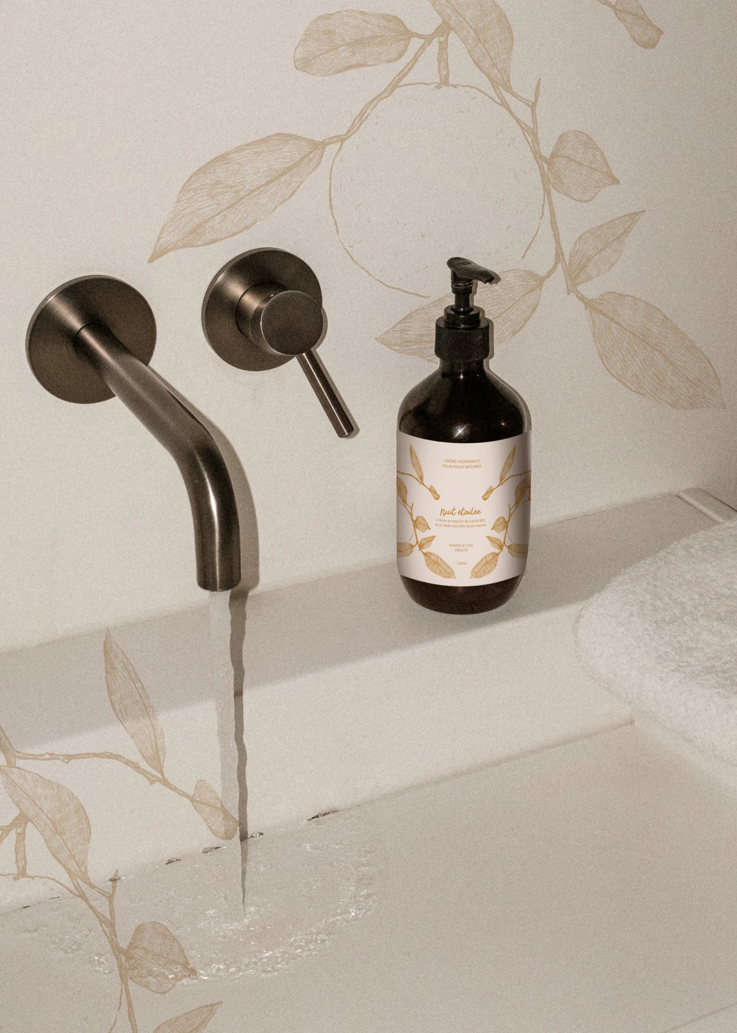 Un lavabo avec un robinet, une bouteille de lotion ou savon et un tapis blanc à proximité dans une salle de bain. Le mur a un motif floral beige.