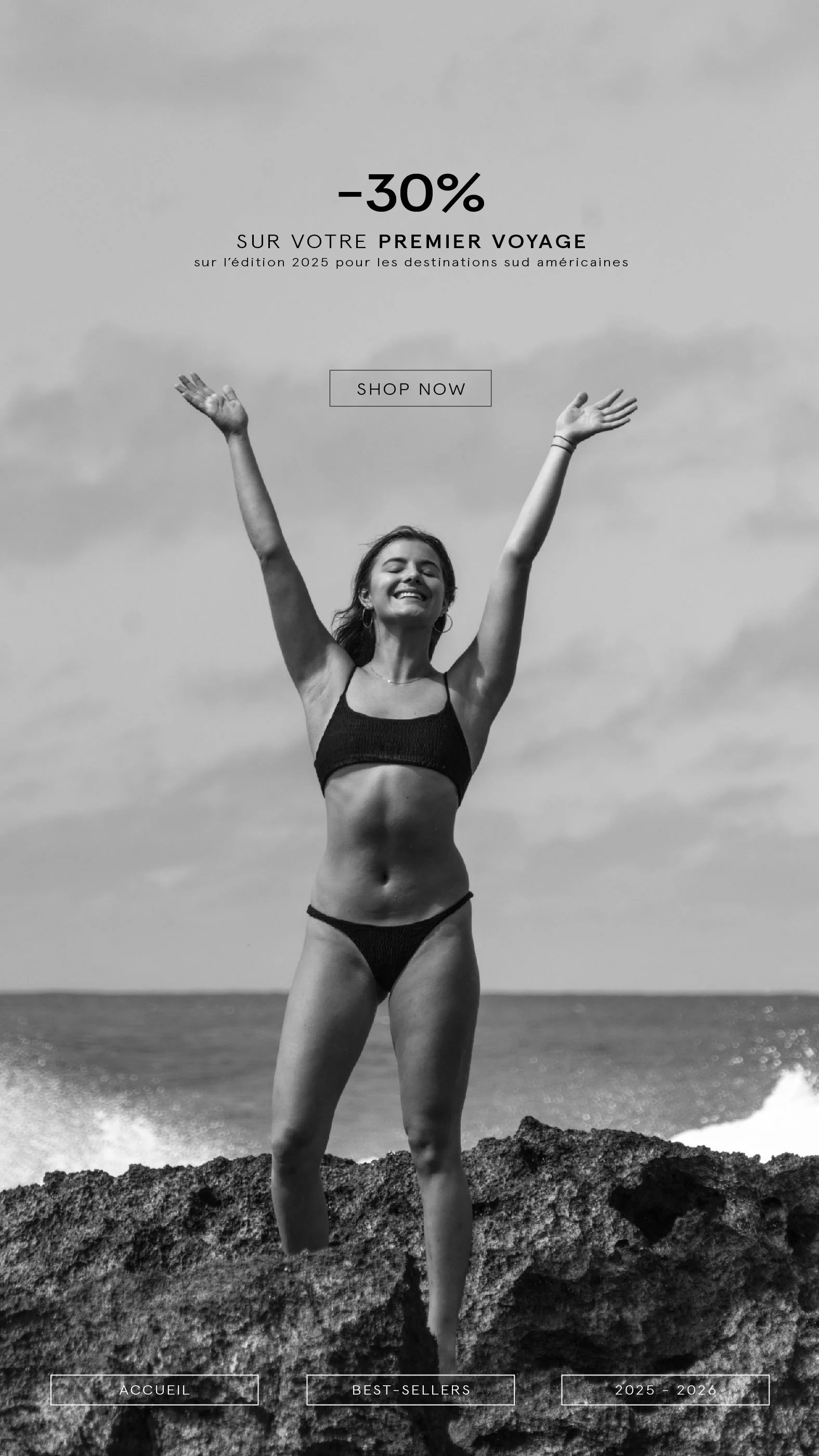 Une femme souriante en maillot de bain noire, se tenant debout sur des rochers au bord de la mer, bras levés, ciel nuageux en arrière-plan.