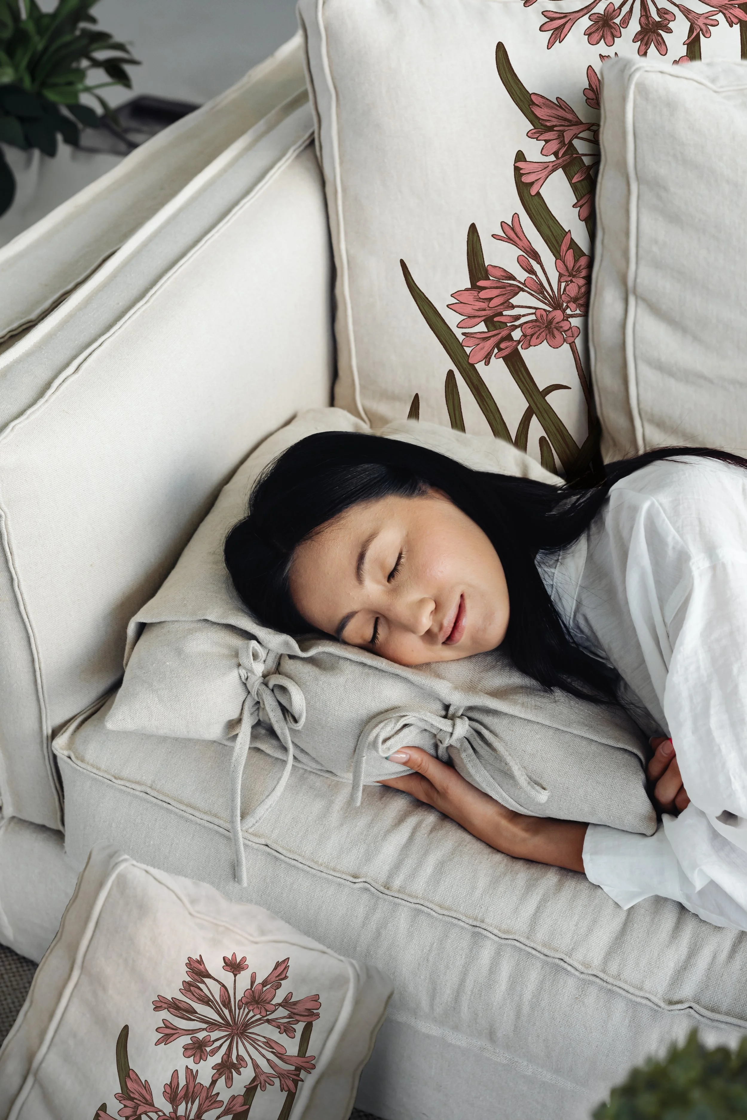 Une femme avec des cheveux noirs dort confortablement sur un canapé beige, entourée de coussins et d'oreillers à motifs floraux roses et verts.