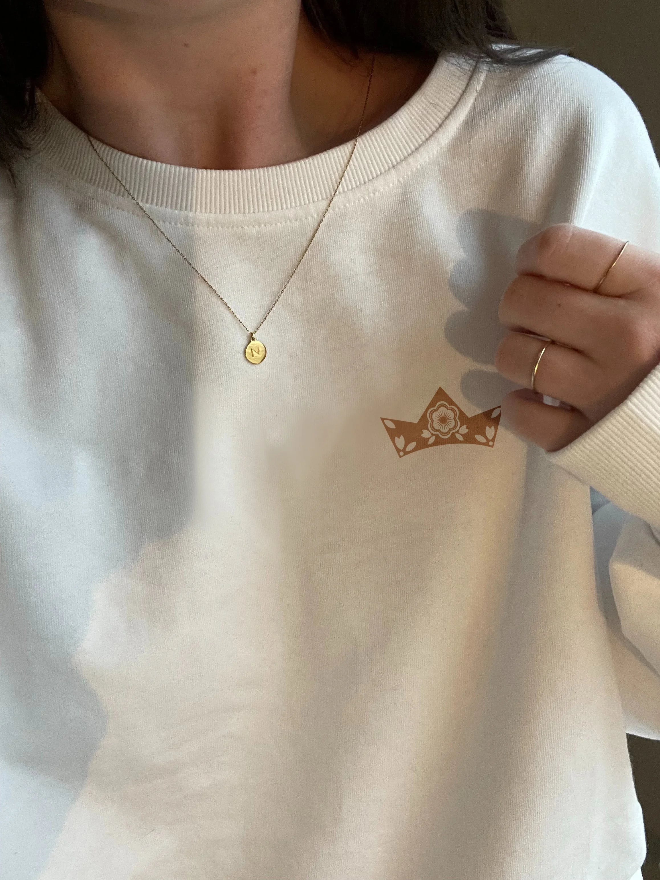Personne portant un sweat blanc avec une petite couronne en dessin sur la poitrine, tenant la manche de son sweat, portant un collier avec un pendentif rond et deux bagues fines aux doigts.