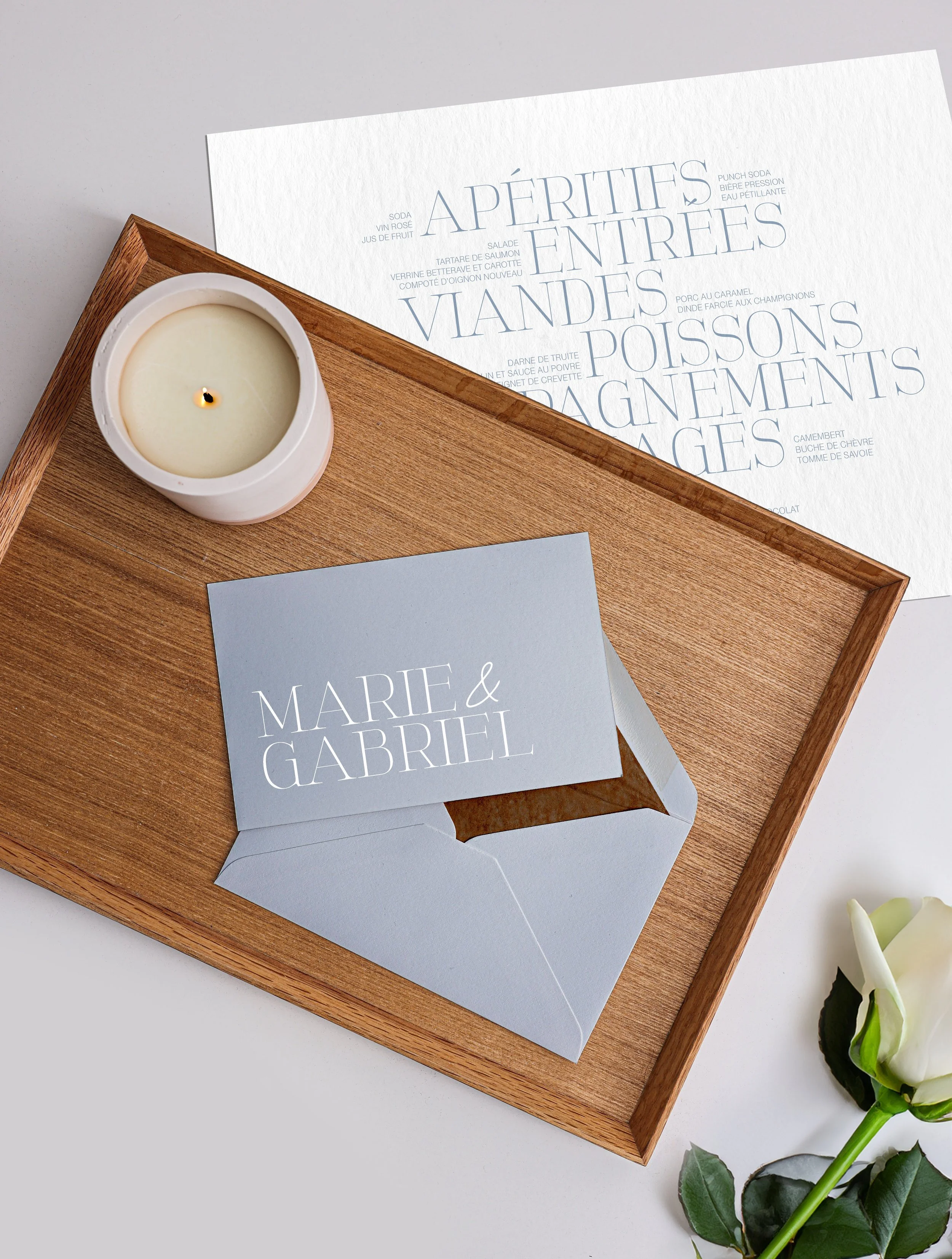 Une planche en bois avec une bougie, une carte de remerciements bleue avec le nom Marie & Gabriel, et un menu de repas écrit en français.