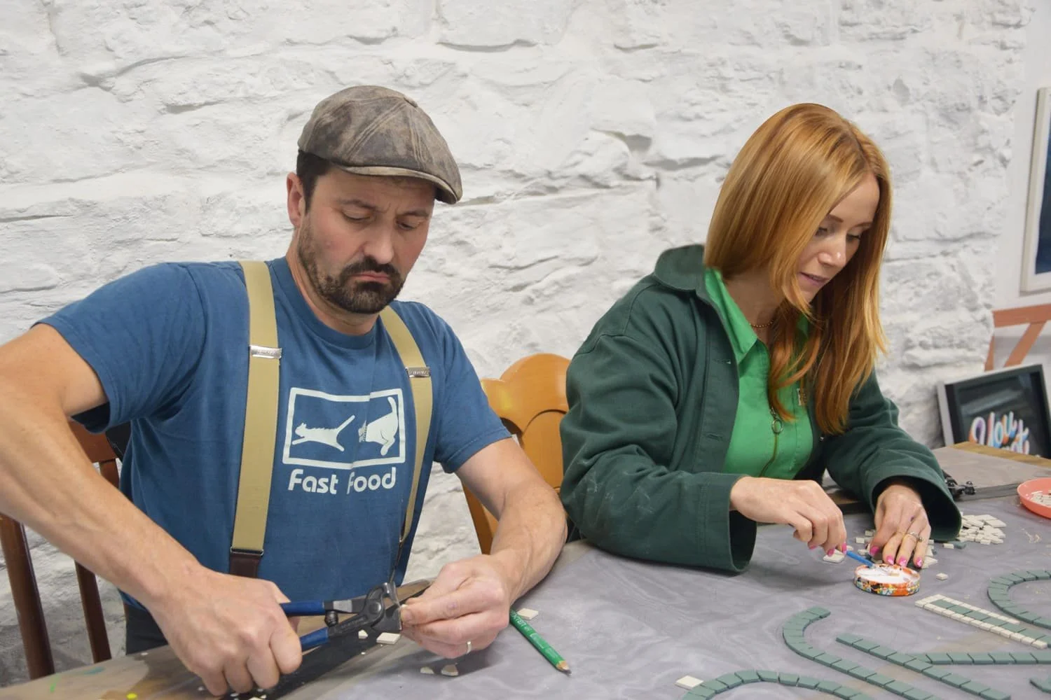 A man with a beard wearing a blue T-shirt with 'Fast food' printed on it, and suspenders, is working with pliers, while a woman with red hair, wearing a green zip-up jacket, is placing small tiles on a table to build a mosaic.