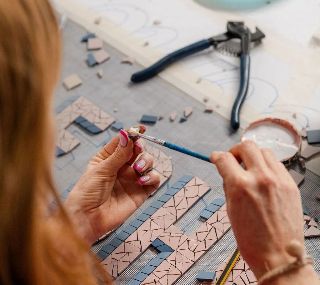 A person working on mosaic tiles, applying grout with a brush at a worktable with tile pieces and tools.