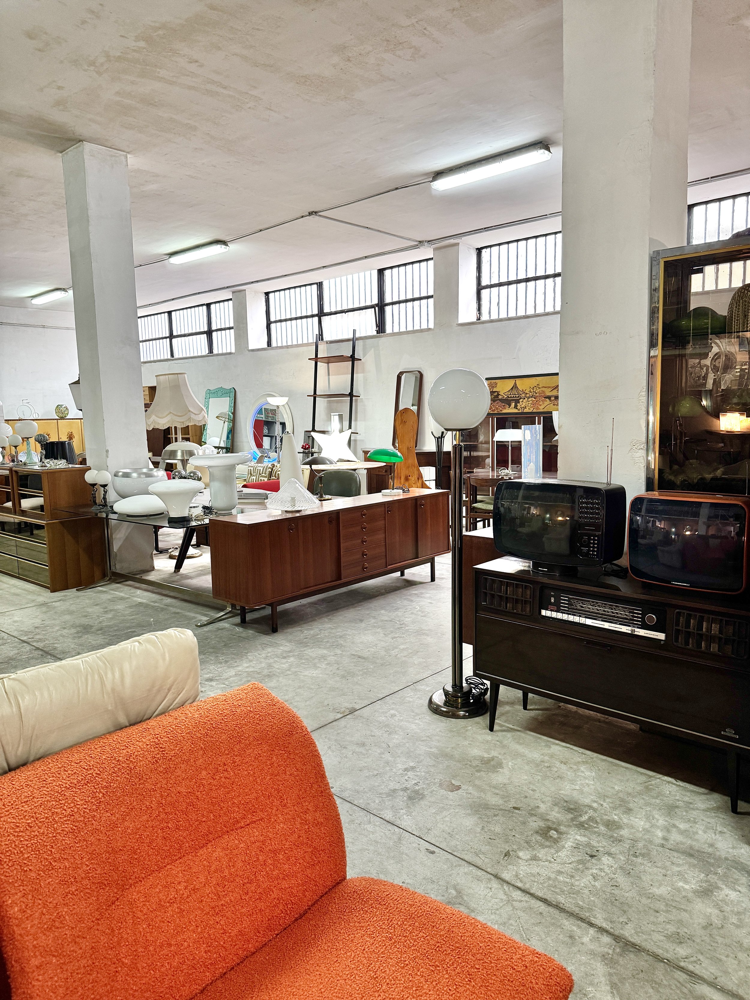 Interno di un negozio di mobili con vari oggetti di arredamento vintage e moderni, tra cui lampade, specchi, sedie e televisori, con pareti di colore bianco e finestre alte.