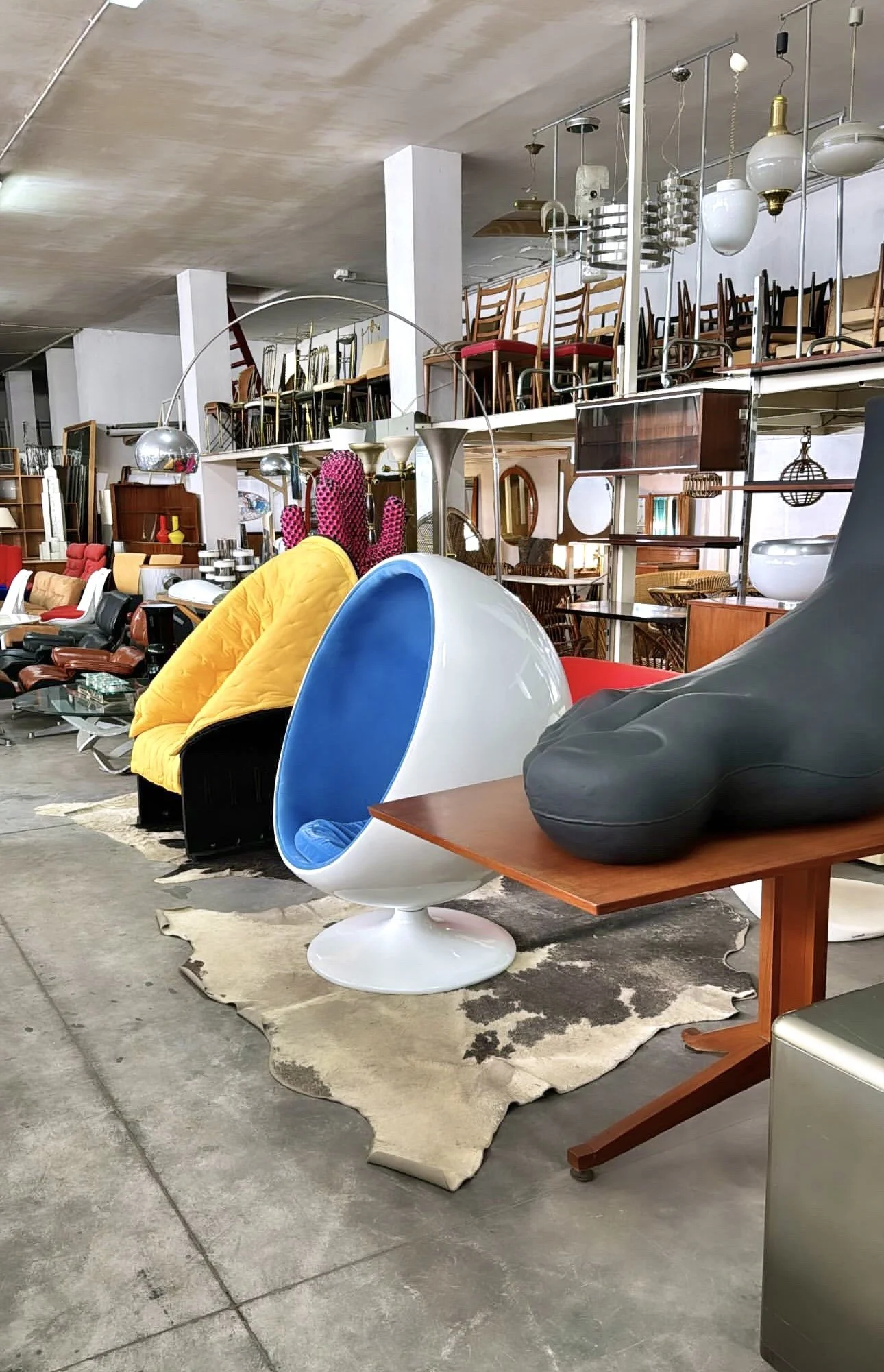 Una vasta selezione di mobili vintage e moderni in un negozio di arredamento, tra cui sedie colorate, lampade e altri oggetti decorativi. In primo piano, ci sono poltrone gialle, un'icona sfera bianca con interno blu, e un grande cuscino grigio su un tavolino di legno. Sul pavimento si vede una pelle di animale beige e marrone.