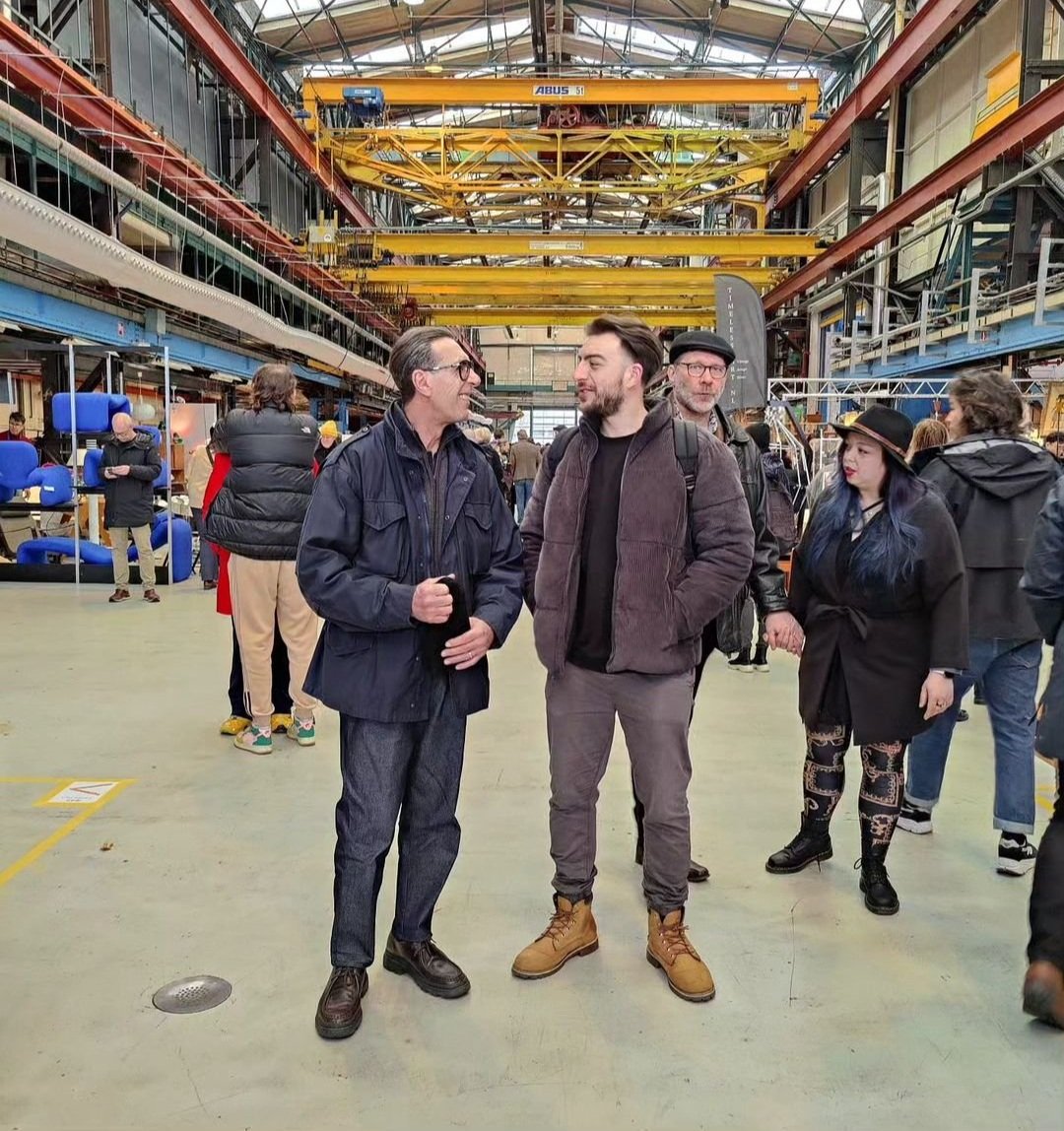 Gruppo di persone che conversano in un'area industriale, probabilmente in una fiera o un evento, con impalcature e gru gialle sul soffitto.