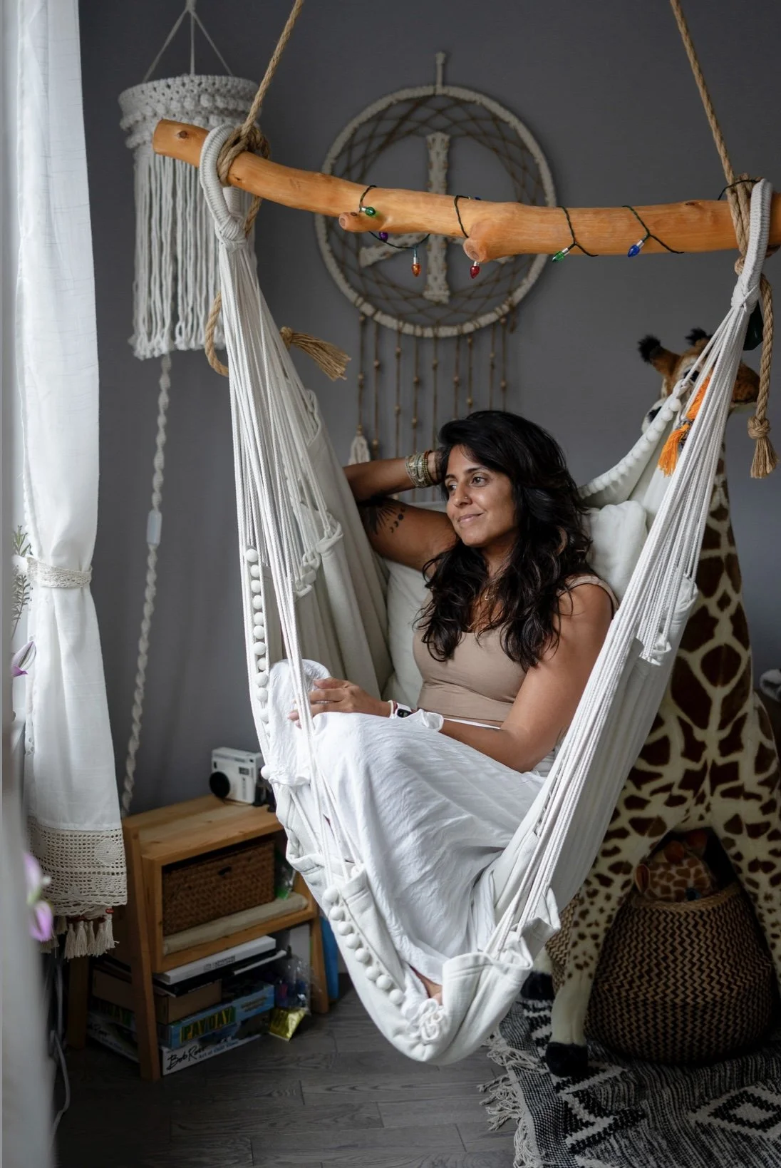 Woman relaxing in a white hammock chair with a giraffe plush toy behind her, in a cozy room decorated with a nautical wall hanging and string lights.