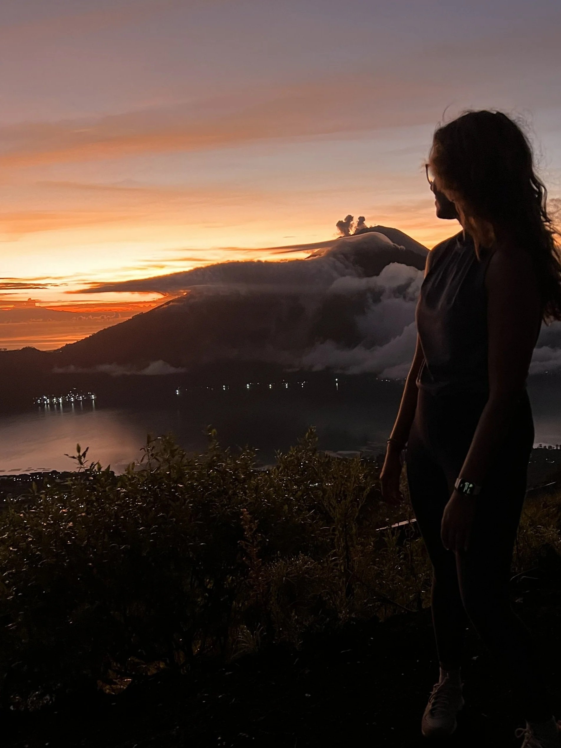 A woman standing outdoors during sunset near a volcanic mountain with smoke and clouds rising from it, overlooking a body of water with lights in the distance.