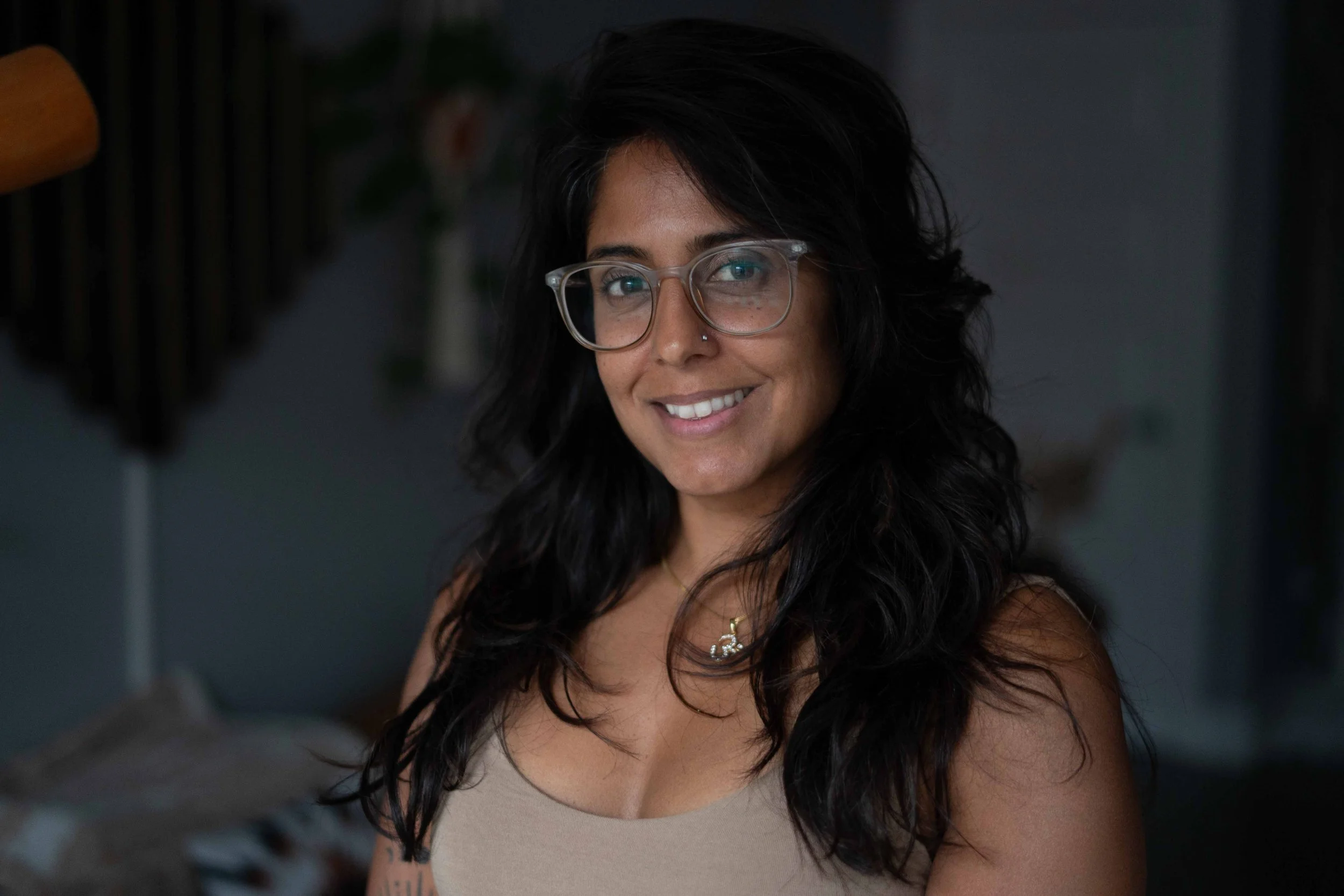 A woman with long dark wavy hair wearing glasses, a beige sleeveless top, and a necklace, smiling indoors with a blurred background.