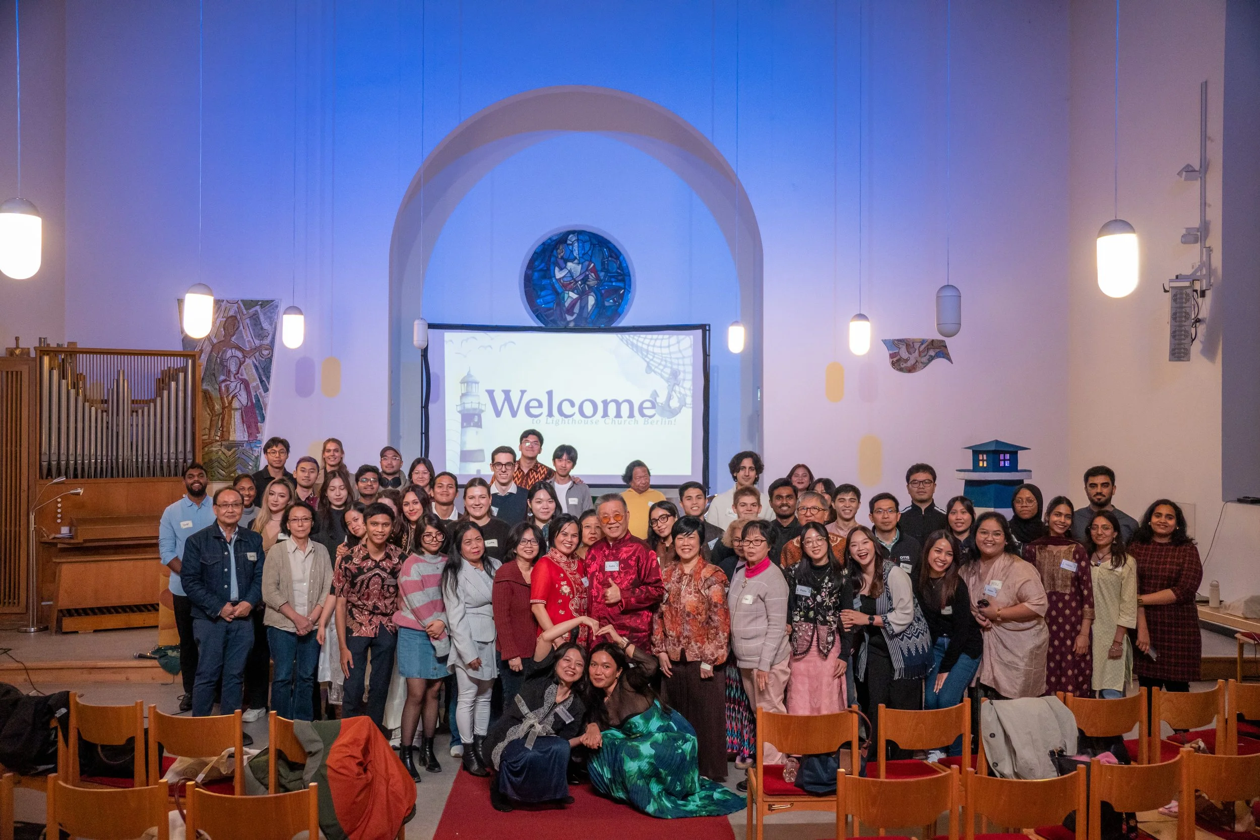 Group of diverse people gathered in a church for a photo, with a 'Welcome' sign on a screen behind them, stained glass window above, and church interior decor.