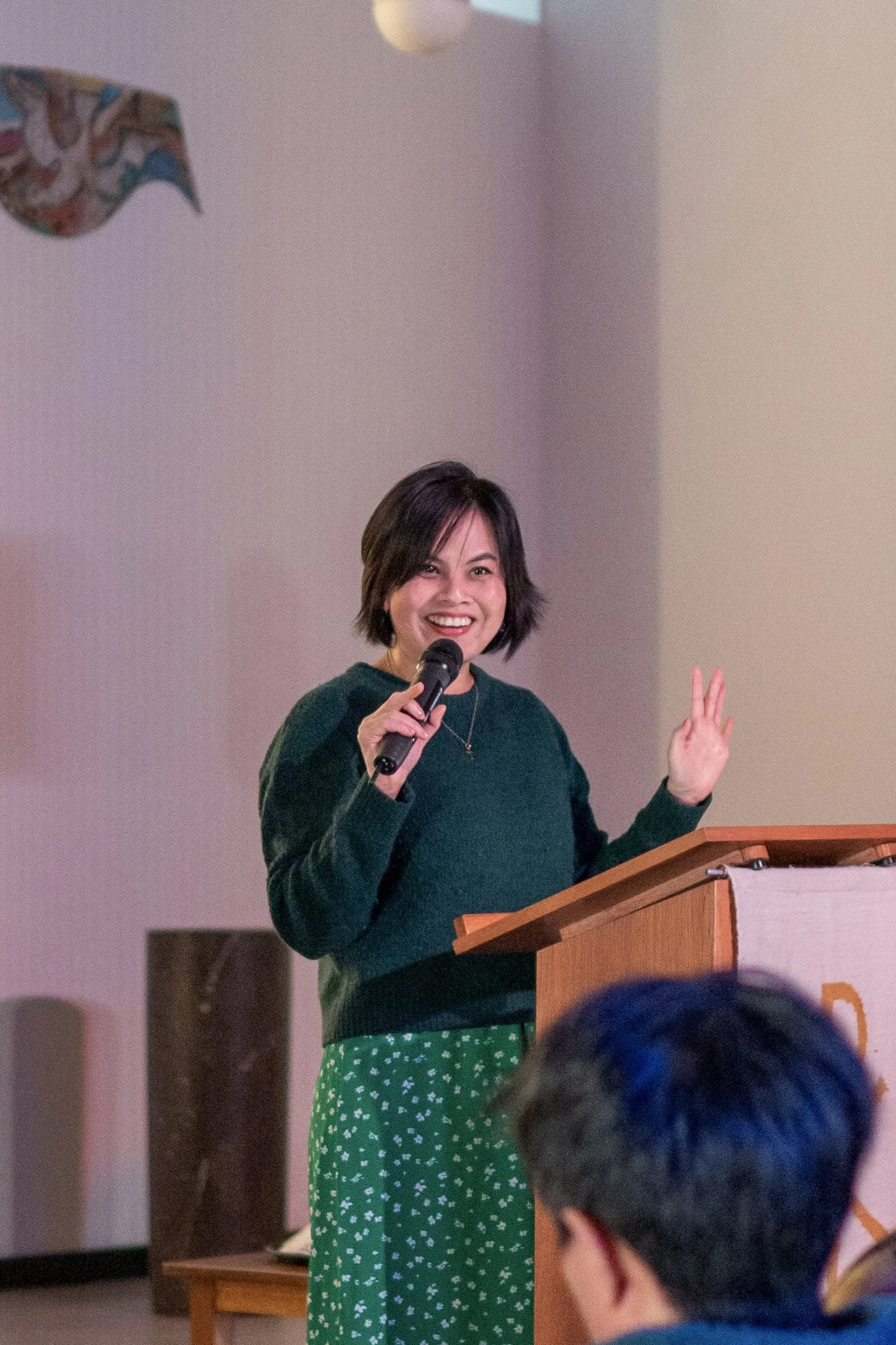 A woman with short dark hair speaking into a microphone at a podium, smiling, and raising her left hand in a welcoming gesture.