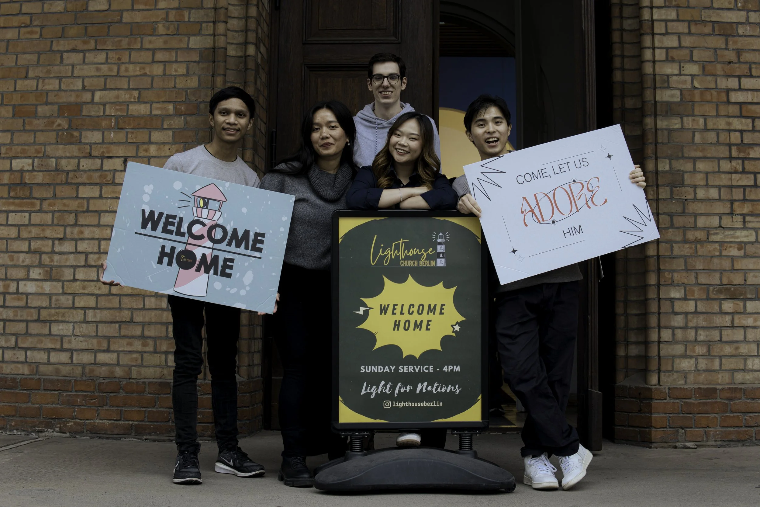 A diverse group of five young adults standing outside a brick church, holding welcome signs and a large welcome message board for a church service event.