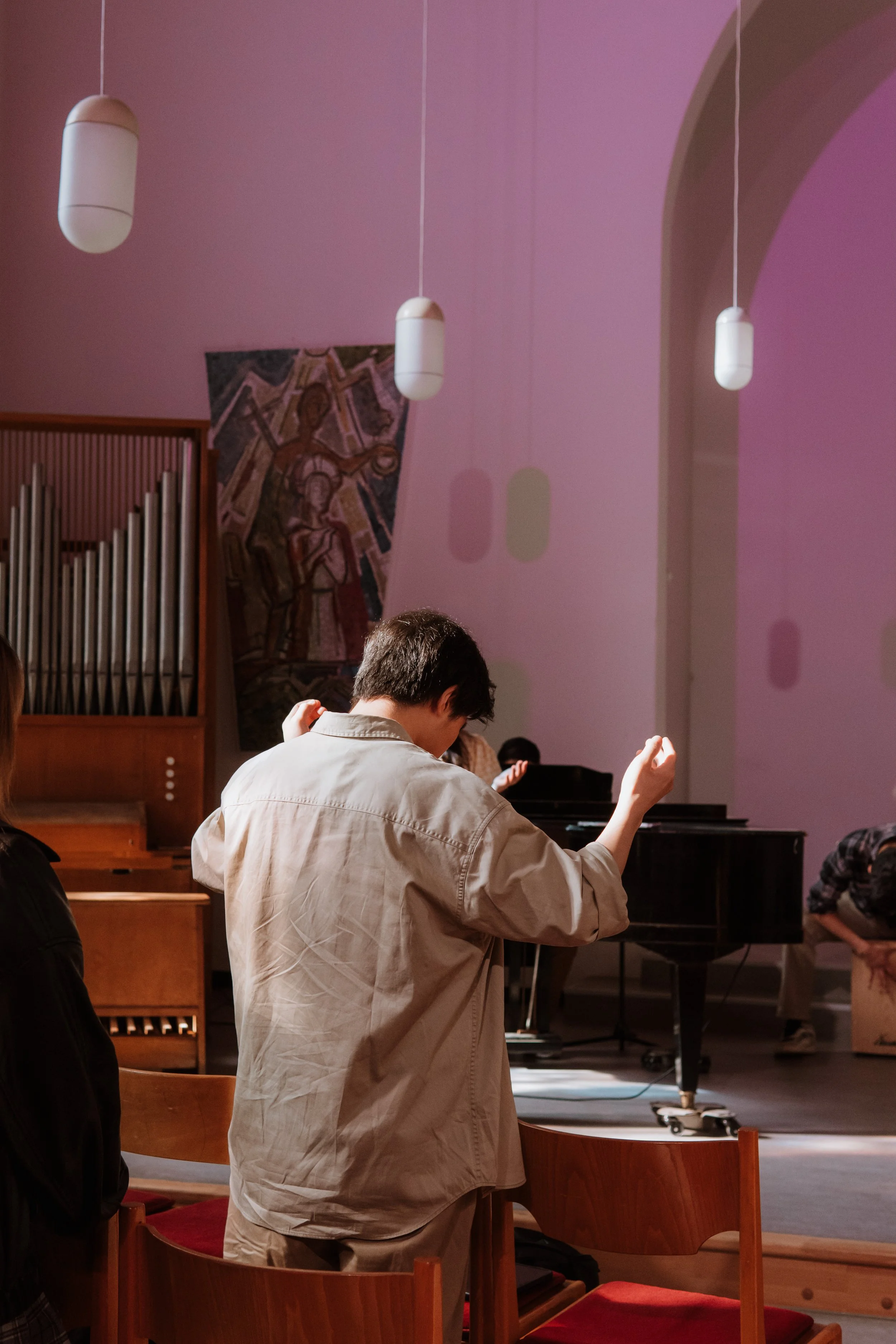 A person in a tan jacket with dark hair is standing in a room with wooden chairs and a piano, raising one arm while facing away from the camera. There are hanging lights and a pink and purple wall with abstract shadows, and religious artwork in the b