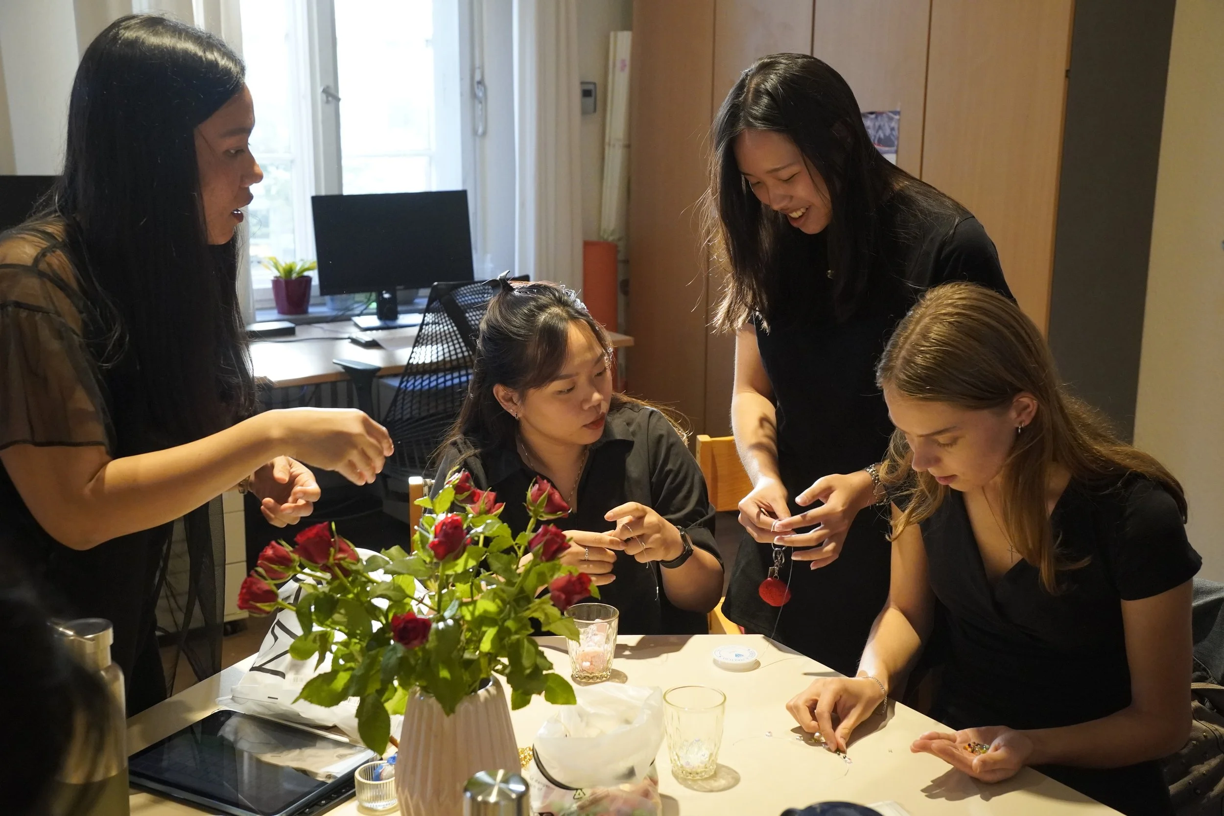Four women gathered around a table with a flower vase, handling jewelry and engaging in conversation.