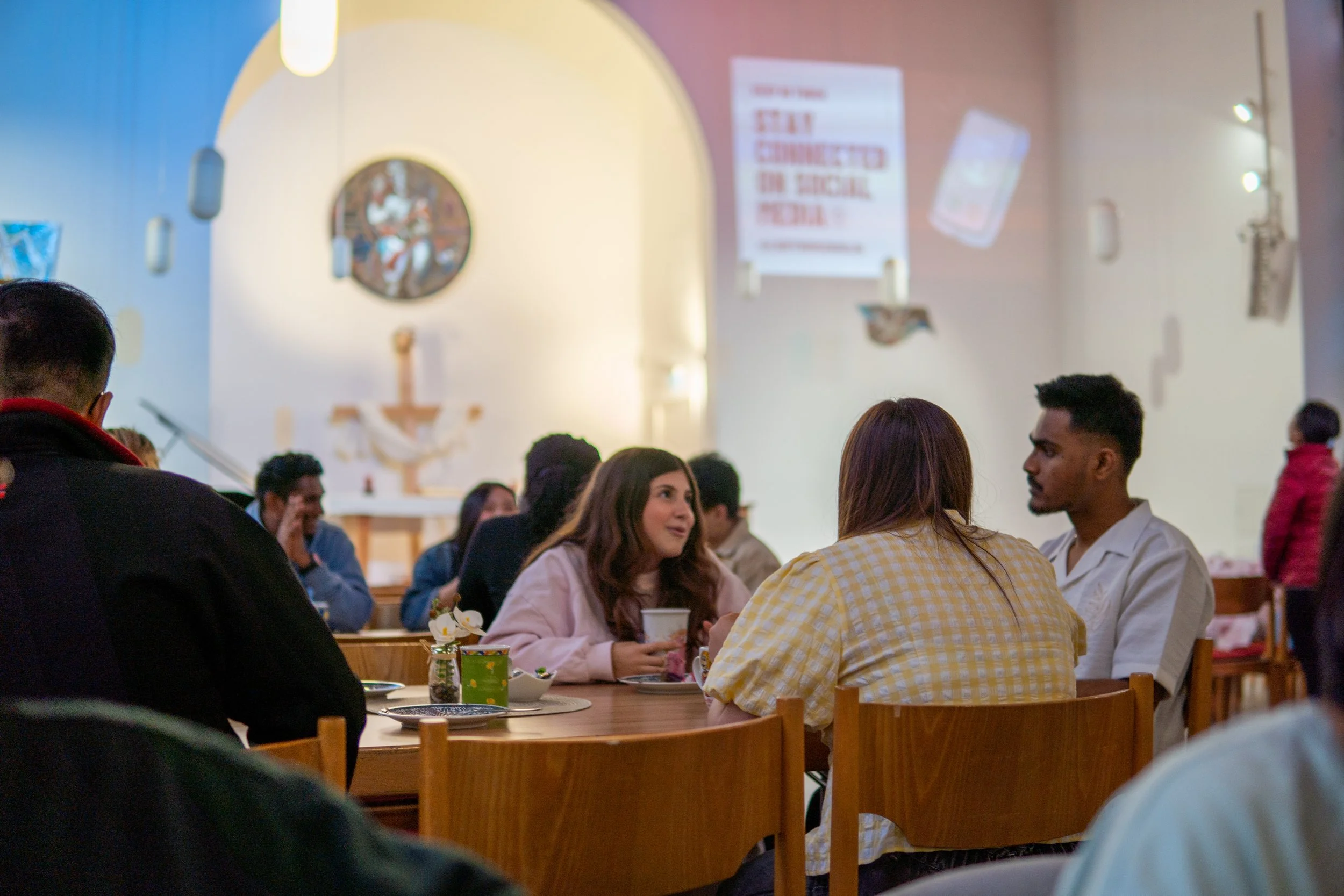 People sitting at a wooden table inside a church, engaged in conversation, with a crucifix on the wall and religious decorations in the background.
