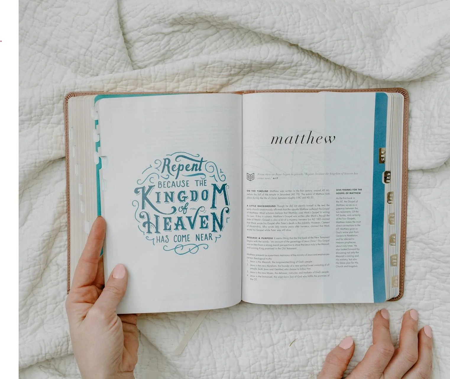 Open Bible on a white bed with one hand holding the left page and the other resting on the bed. The left page features a decorative quote about God's kingdom and heaven, while the right page has the name "Matthew" and an introductory text.